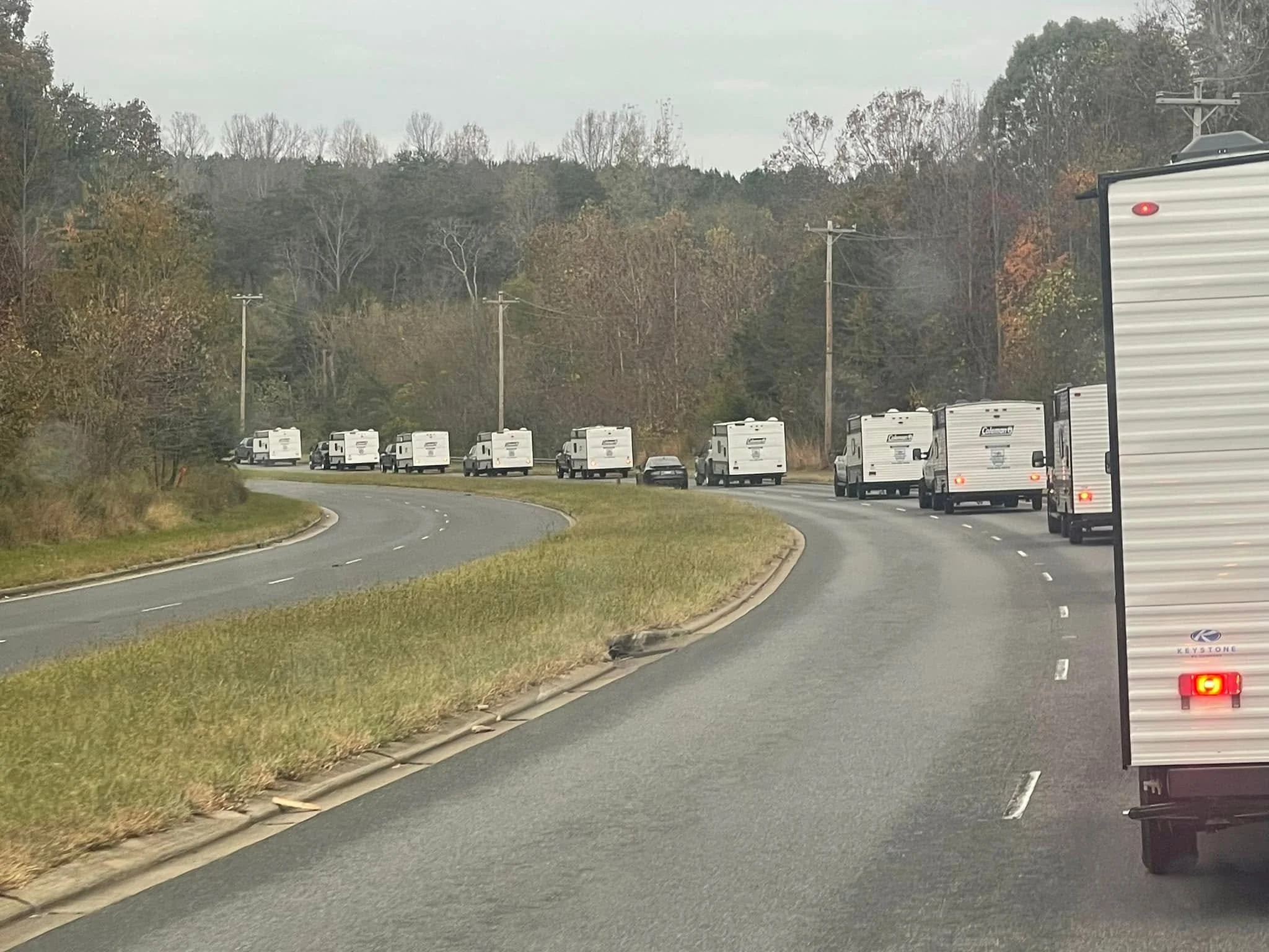 Line of parked travel trailers and fifth-wheel RVs along the side of a curved road, with a wooded hillside in the background.