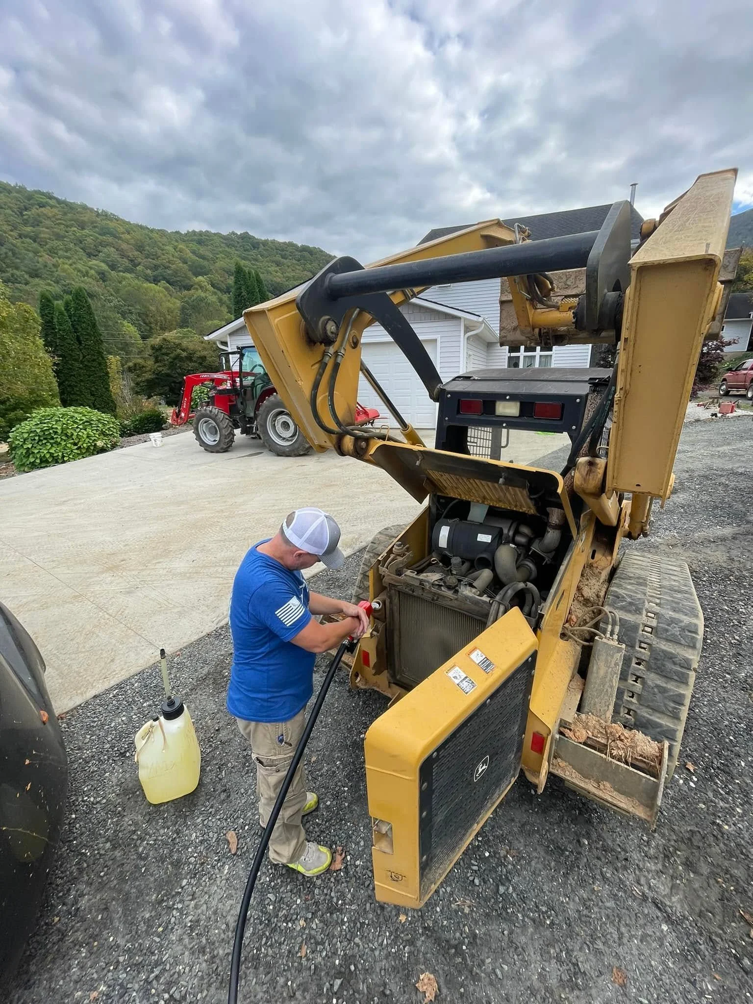 A man in a blue shirt, khakis, and a white cap working on a yellow construction machine in a residential driveway. A red tractor is in the background, with mountains and clouded sky overhead.