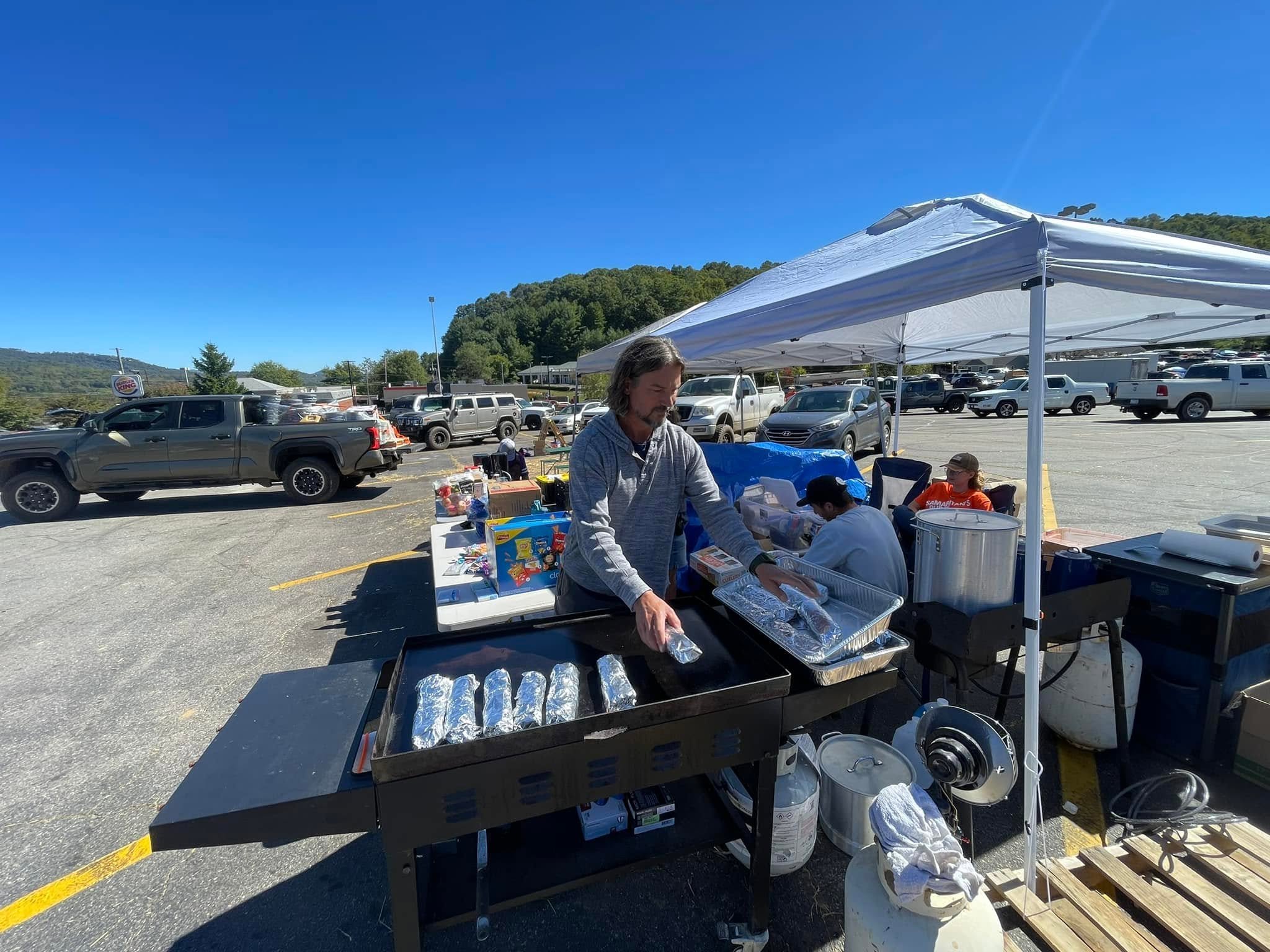 People preparing food at a small outdoor stand in a parking lot, under a white canopy, with parked cars and green hills in the background on a sunny day.