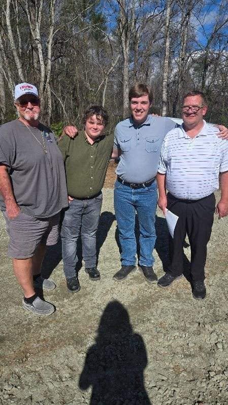 Four men standing outdoors in a wooded area, smiling, with a shadow of a person taking the photo in the foreground.