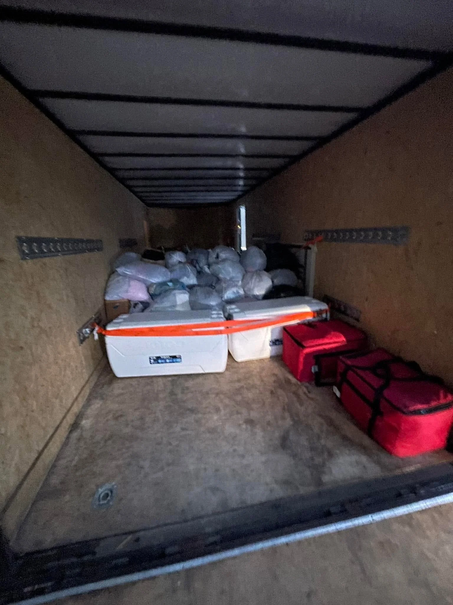 View inside a storage compartment showing bags, a white cooler, and red toolbox, with a metal rack mounted on the side wall.