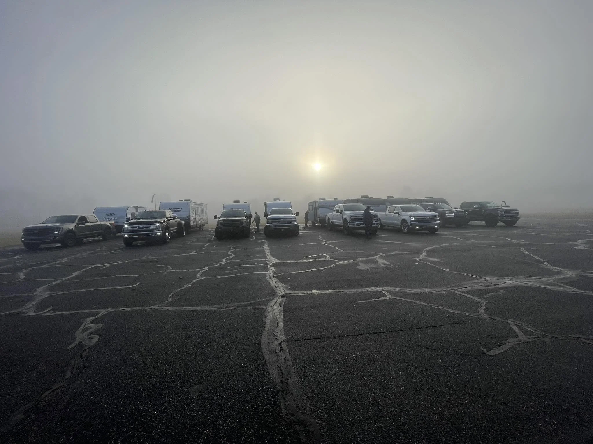 A parking lot with several vehicles, including trucks and RVs, under a foggy sky with visible sunlight.