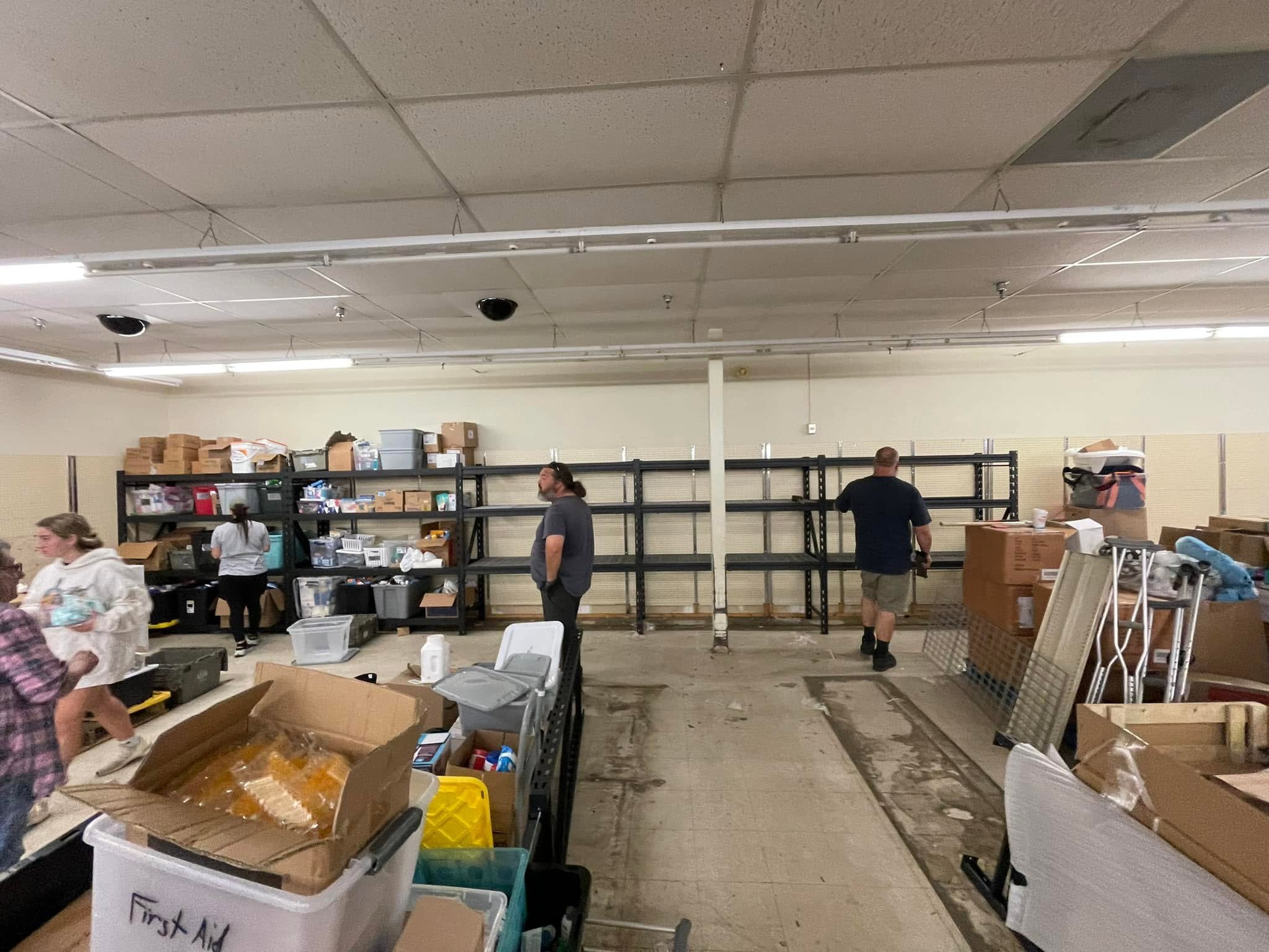 People organizing supplies and boxes in a storage room with metal shelves, boxes, and various items, under a suspended ceiling with lights and security cameras.