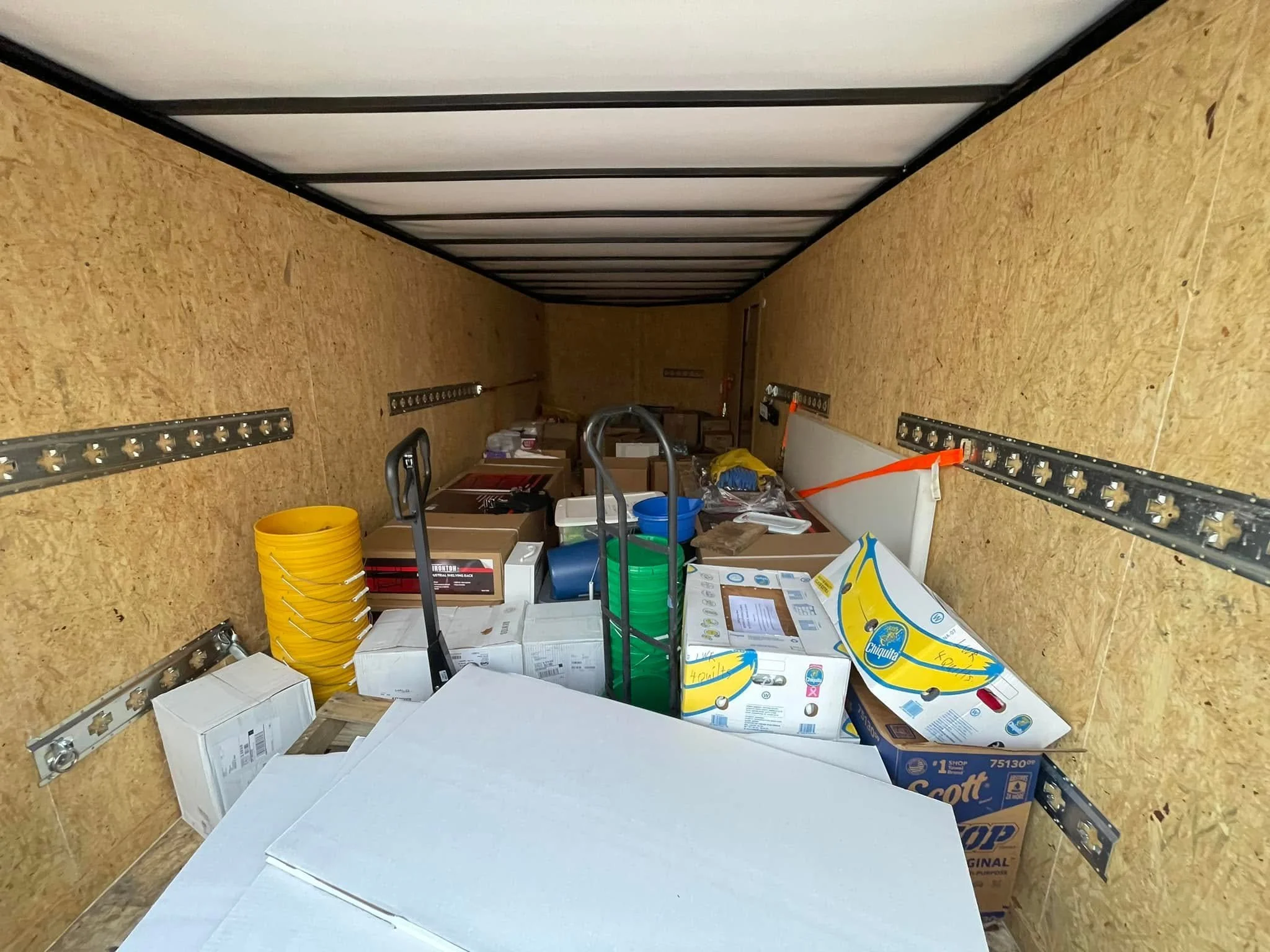 A storage or utility room with plywood walls and ceiling, filled with boxes, buckets, and tools, some shelves running along the walls.
