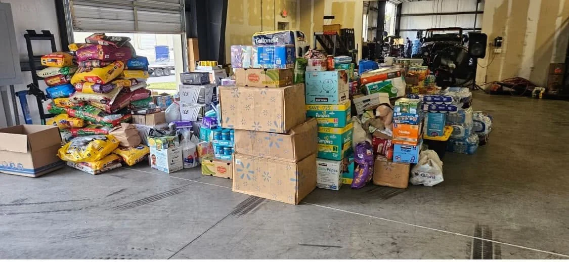 Shelves and stacks of canned and packaged food items, baby diapers, and bottled water in a warehouse.