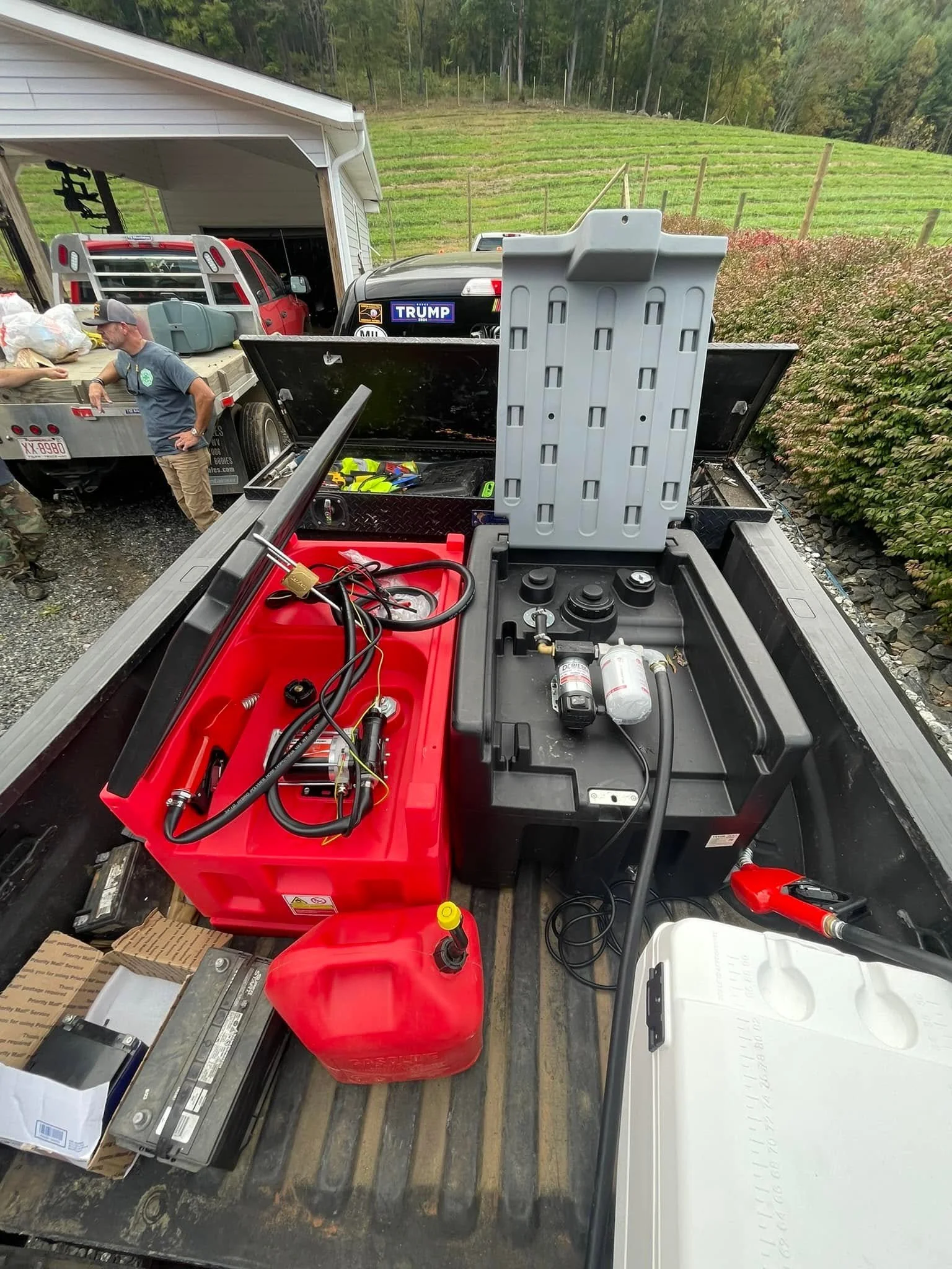 Pickup truck bed filled with various tools, batteries, and equipment, with a white cooler on the right side and a red fuel container in the center, outside a garage and rural landscape in the background.