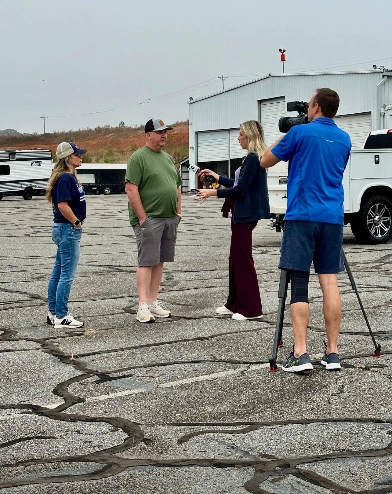 A news crew interviews a man outdoors in a parking lot with a gray building, a white truck, and a television camera crew.