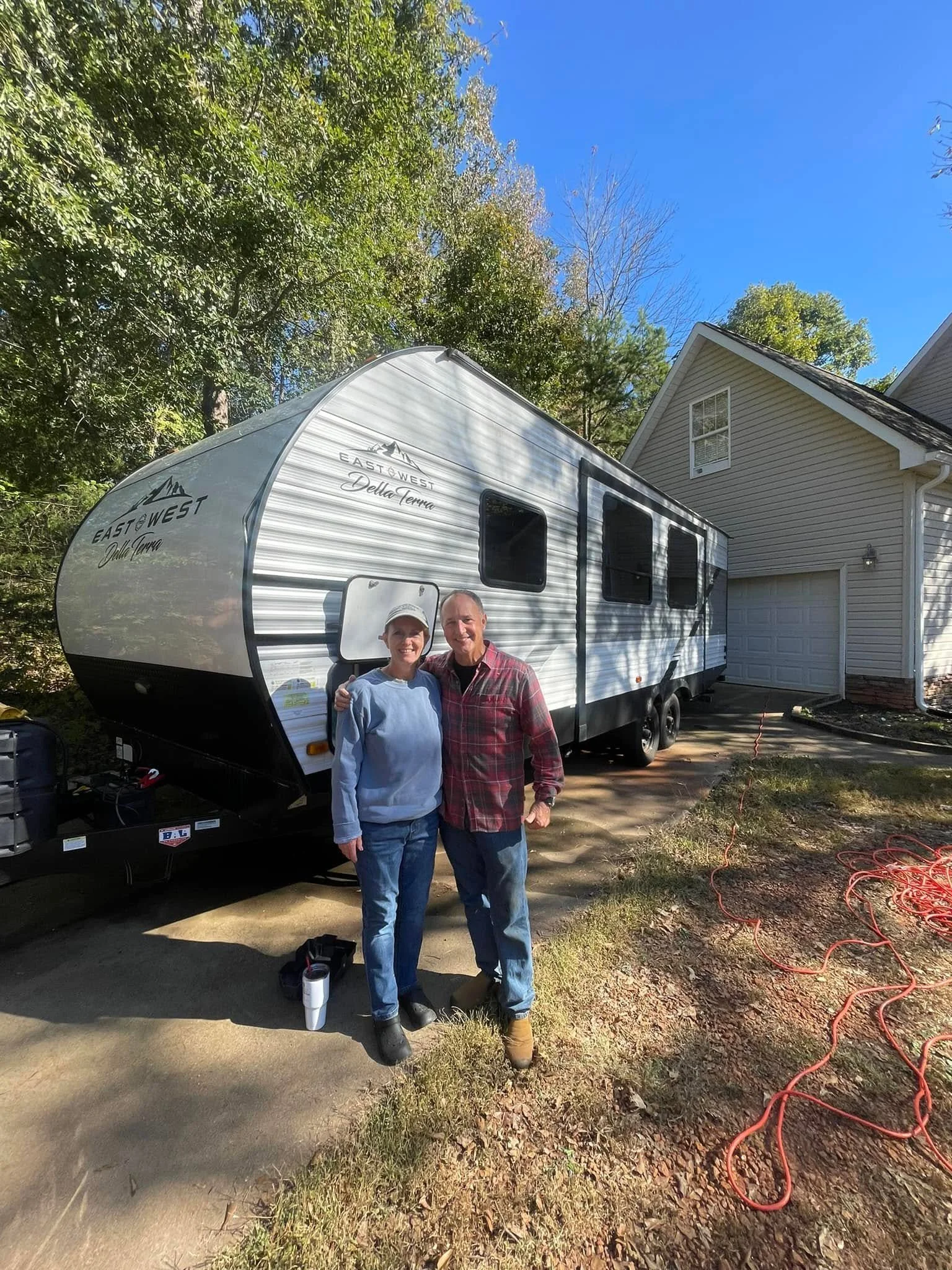 A man and a woman standing outside in front of a travel trailer parked next to a house, with trees and a clear blue sky in the background.