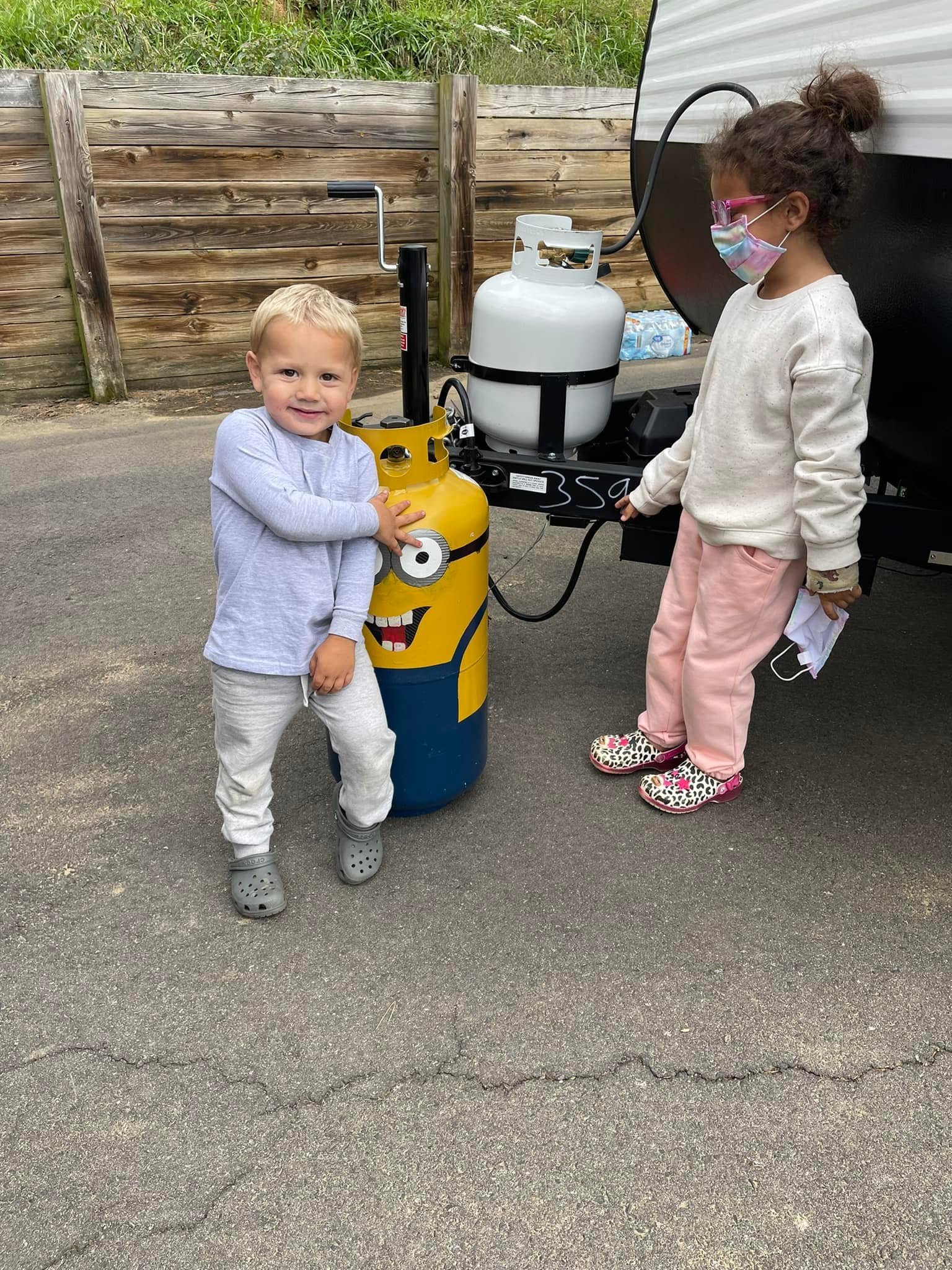Two children near propane tanks, one smiling girl in gray with Crocs, one girl with face mask and glasses wearing pink pants and leopard print shoes, on an asphalt surface with a wooden fence background.