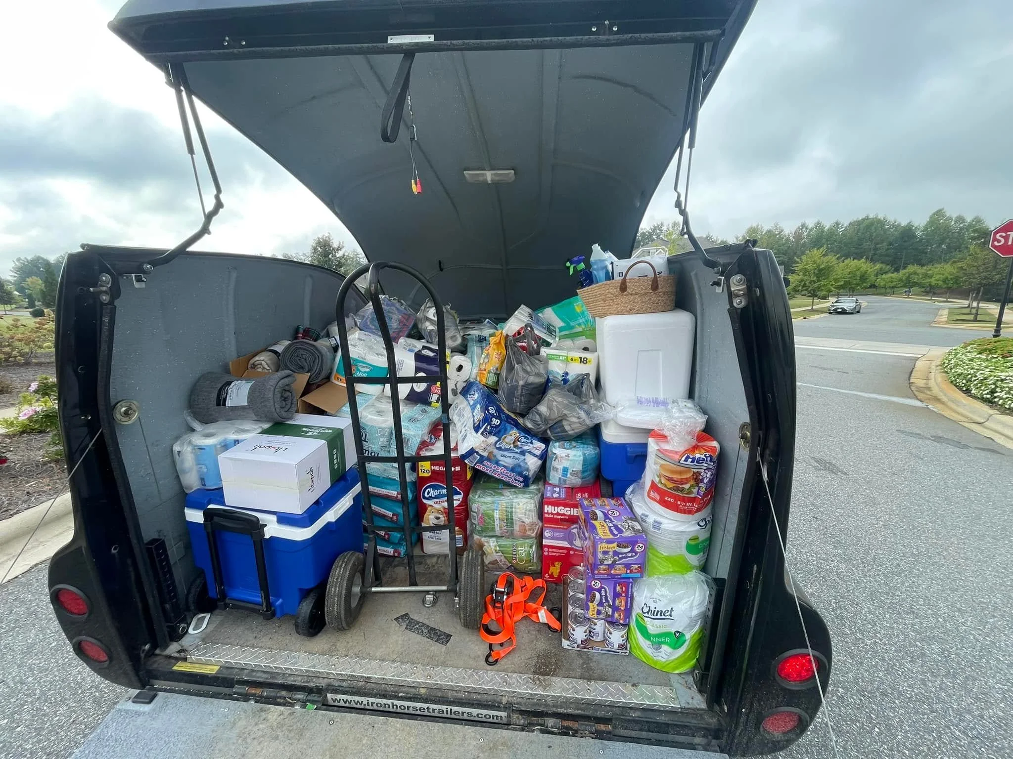 Open trailer packed with groceries including paper towels, bottled water, pet food, and household supplies in a parking lot on a cloudy day.