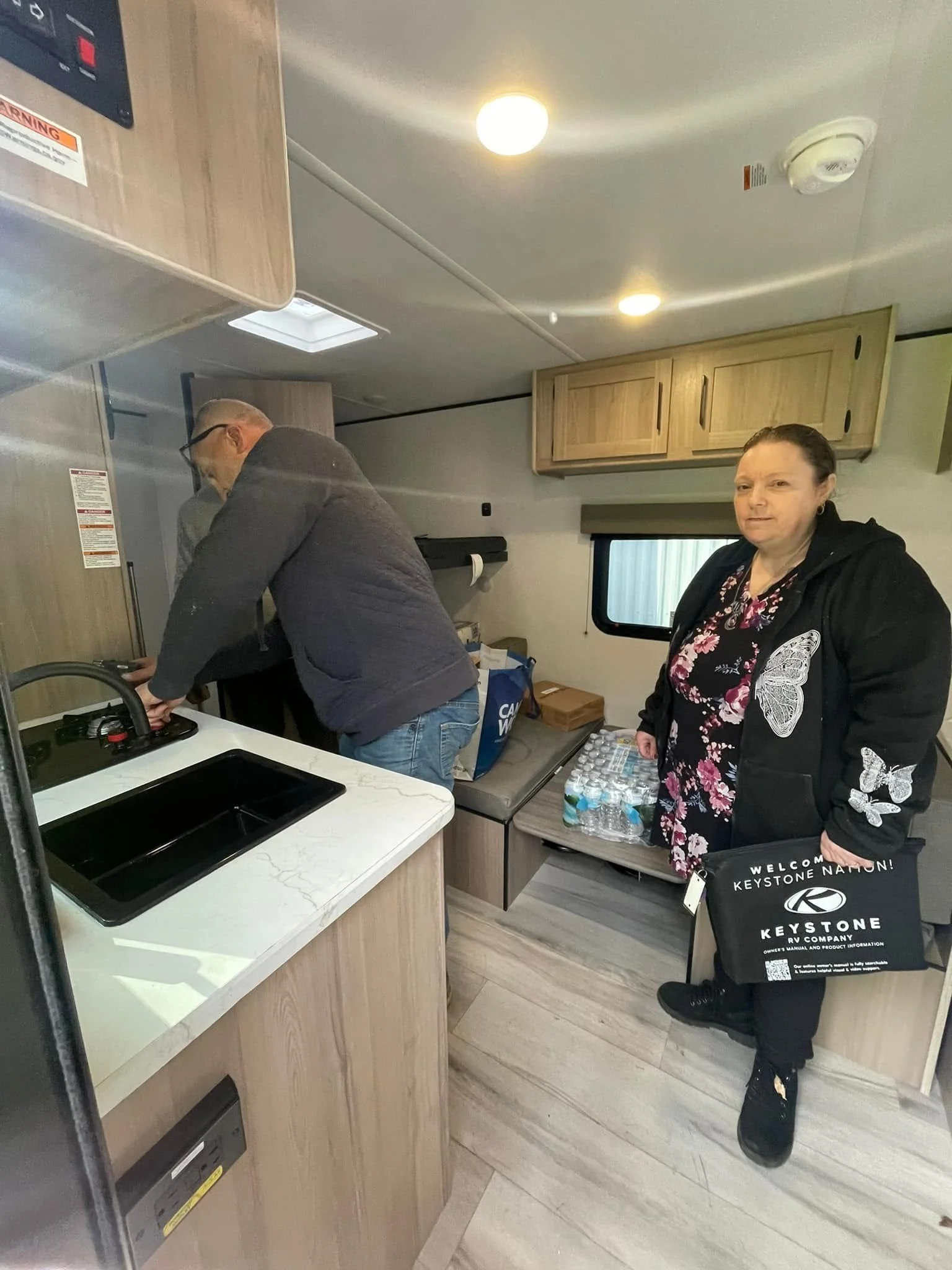Interior of a recreational vehicle with a woman standing near bottled water and a man working at the kitchen sink, wooden cabinets overhead, window with blinds, and ceiling lights.