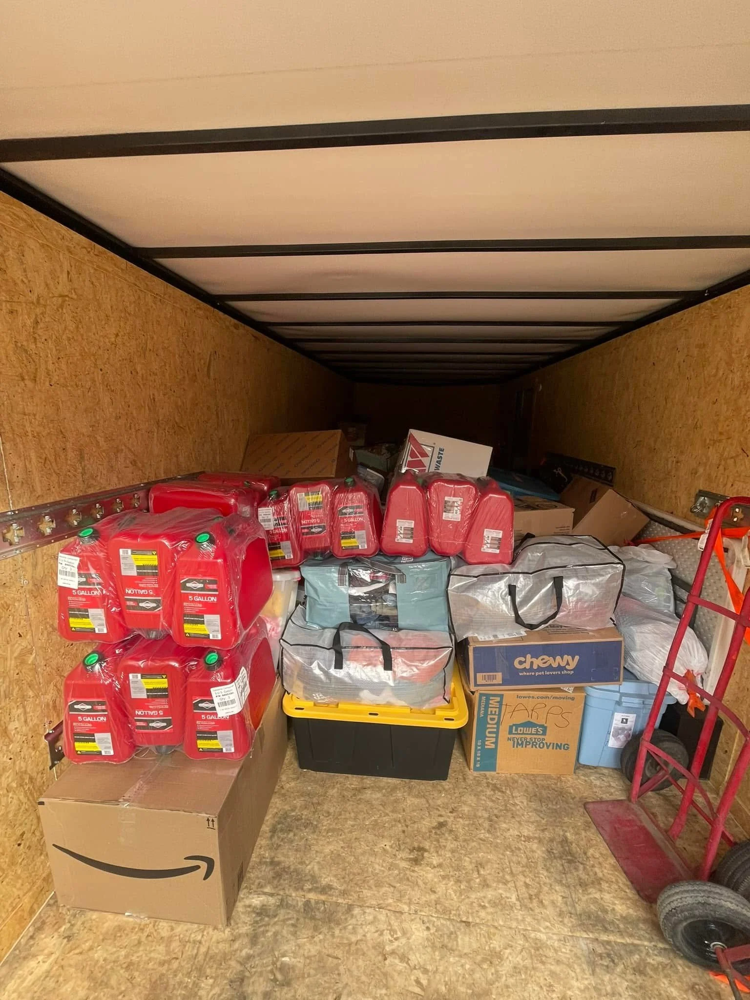 A storage room with 15-gallon containers of red liquid, boxes, bags, and various household items stacked on the floor and against the walls, with a hand truck and a large cardboard box.