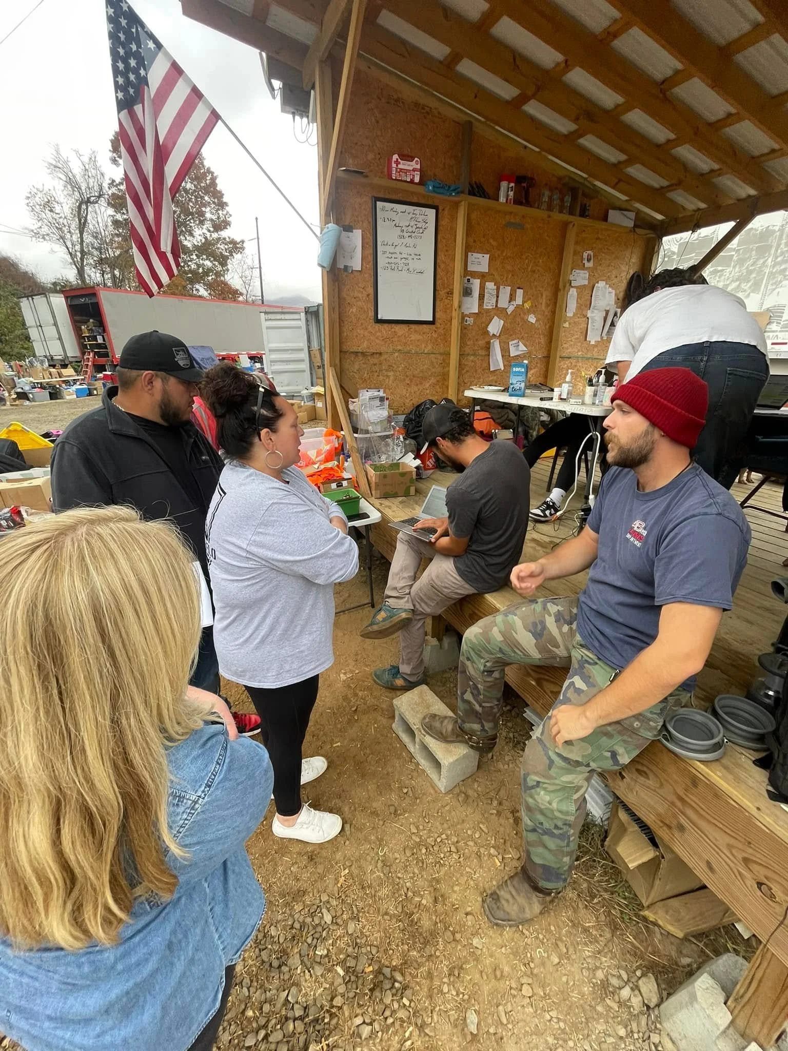 People gathered inside a wooden structure, some sitting and some standing, with a man in camouflage pants and a gray T-shirt sitting on a wooden bench. A woman with a bun and hoop earrings is standing nearby, and another man with a cap is standing an