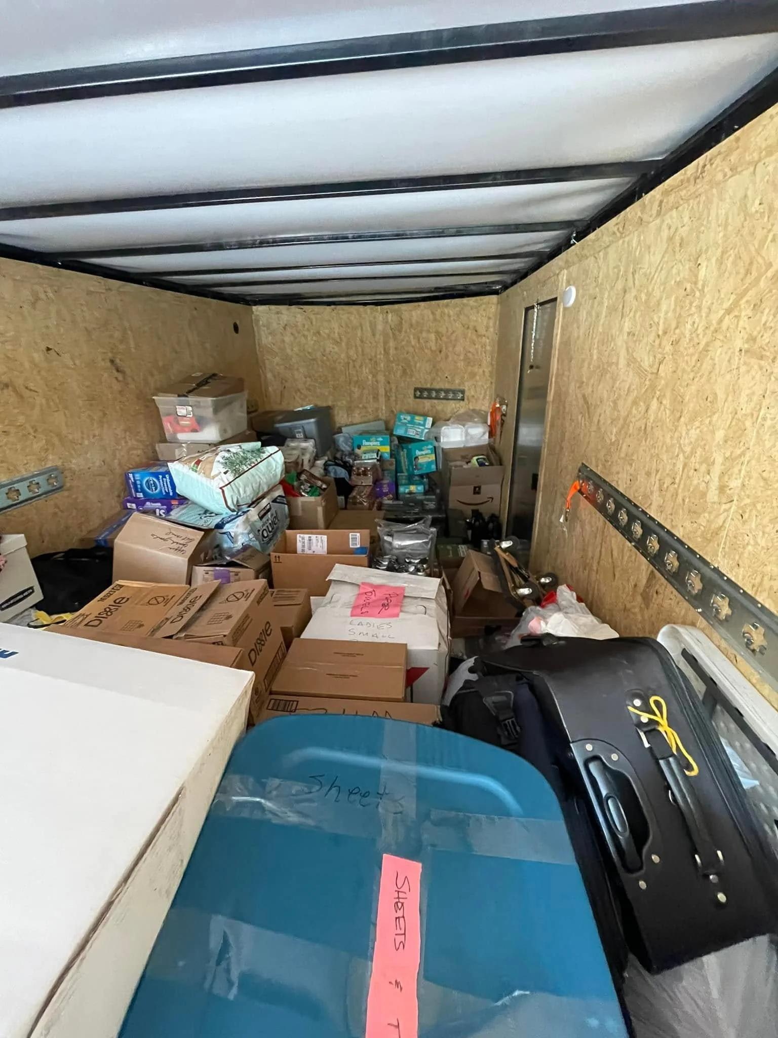 The image shows the inside of a storage or utility trailer with various boxes, bags, and miscellaneous items, including a black toolbox, a pack of paper towels, and a back of an electric grill or cooker. There's a pink labeled sticky note on a blue i