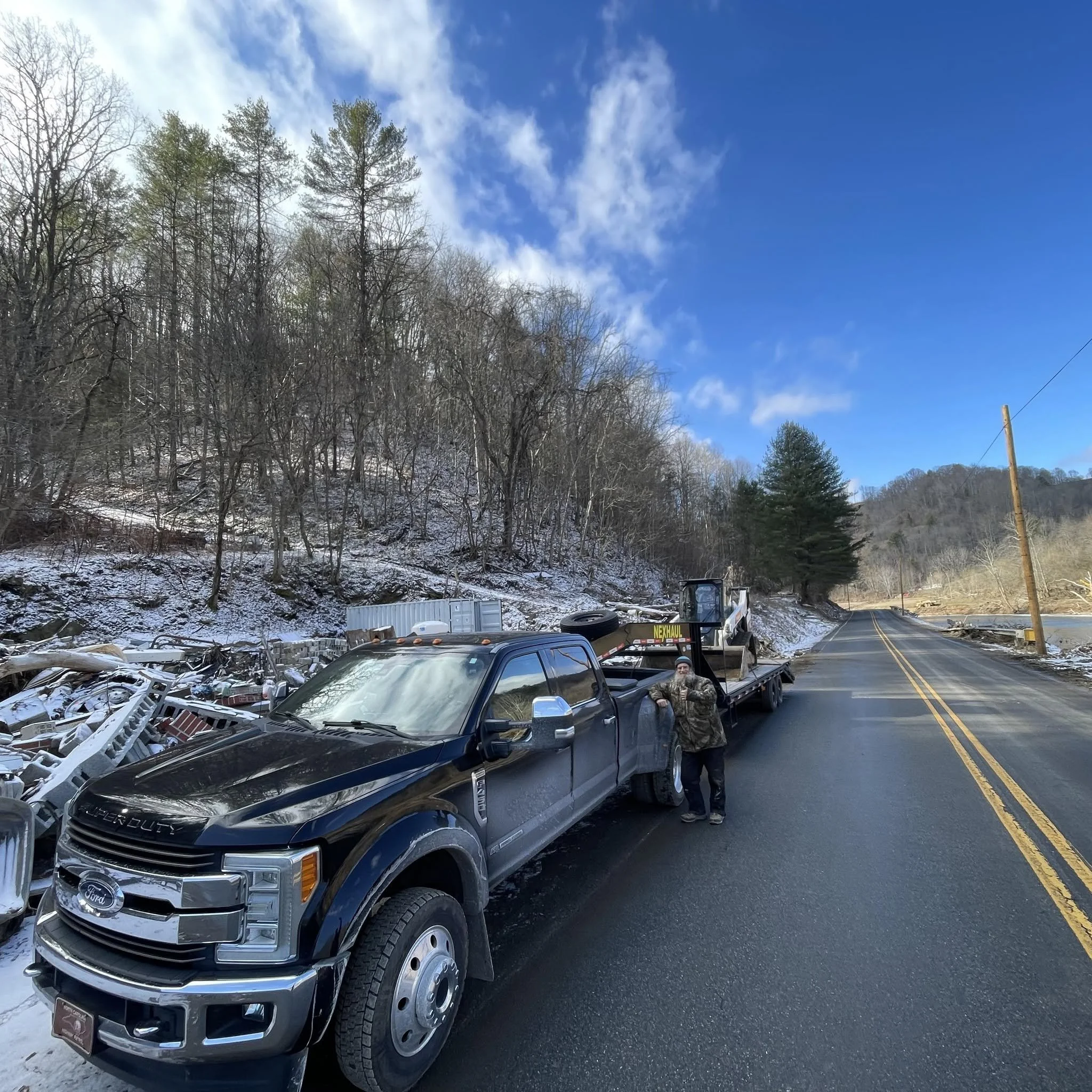 Pickup truck carrying yard debris, parked on the side of a two-lane road with a wooded hillside in the background, under a partly cloudy sky.