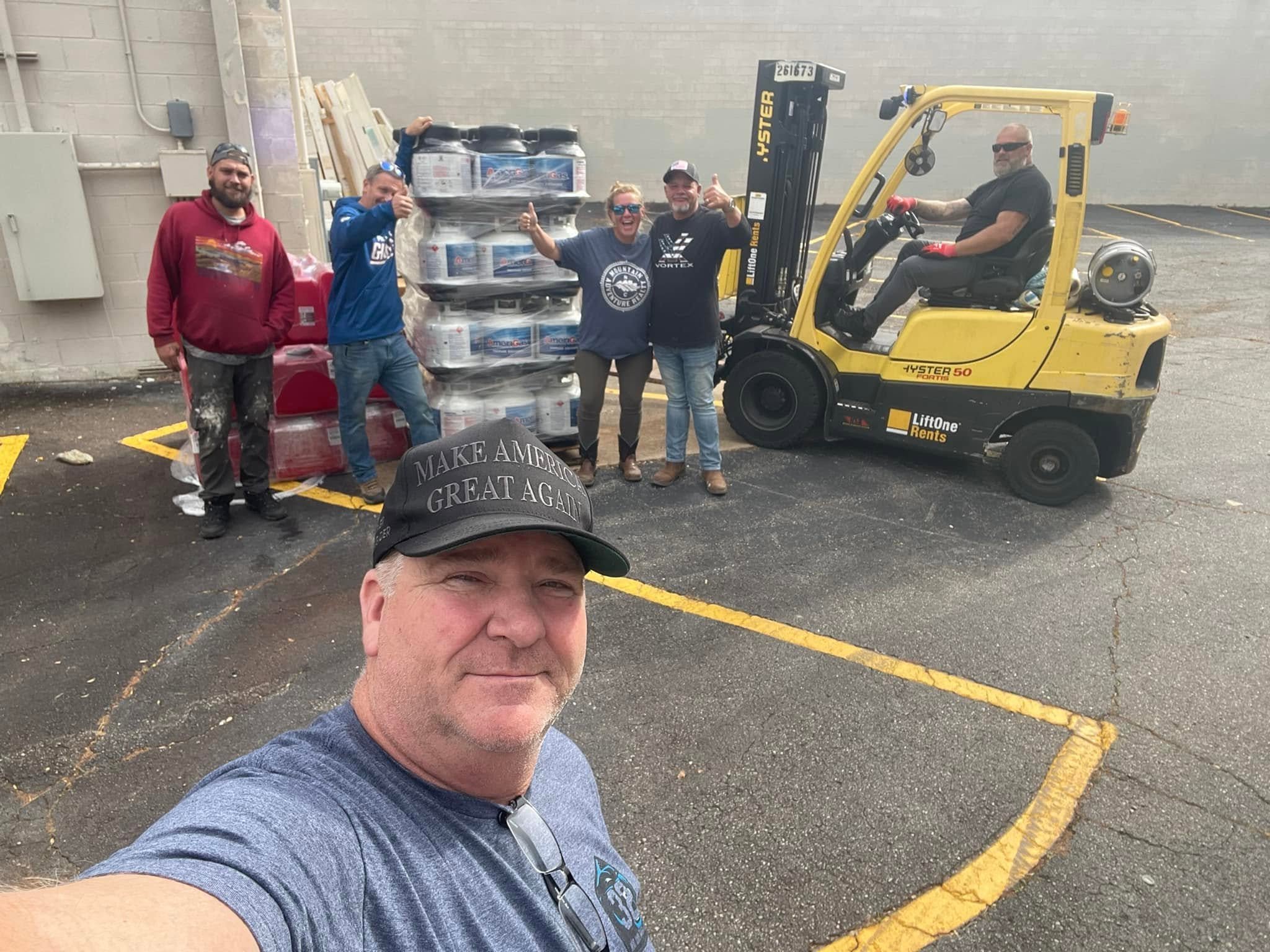 Group of five people standing next to a yellow forklift truck in a parking lot, posing for a selfie. Four of them are smiling and giving thumbs-up signs, while one person is sitting on the forklift.