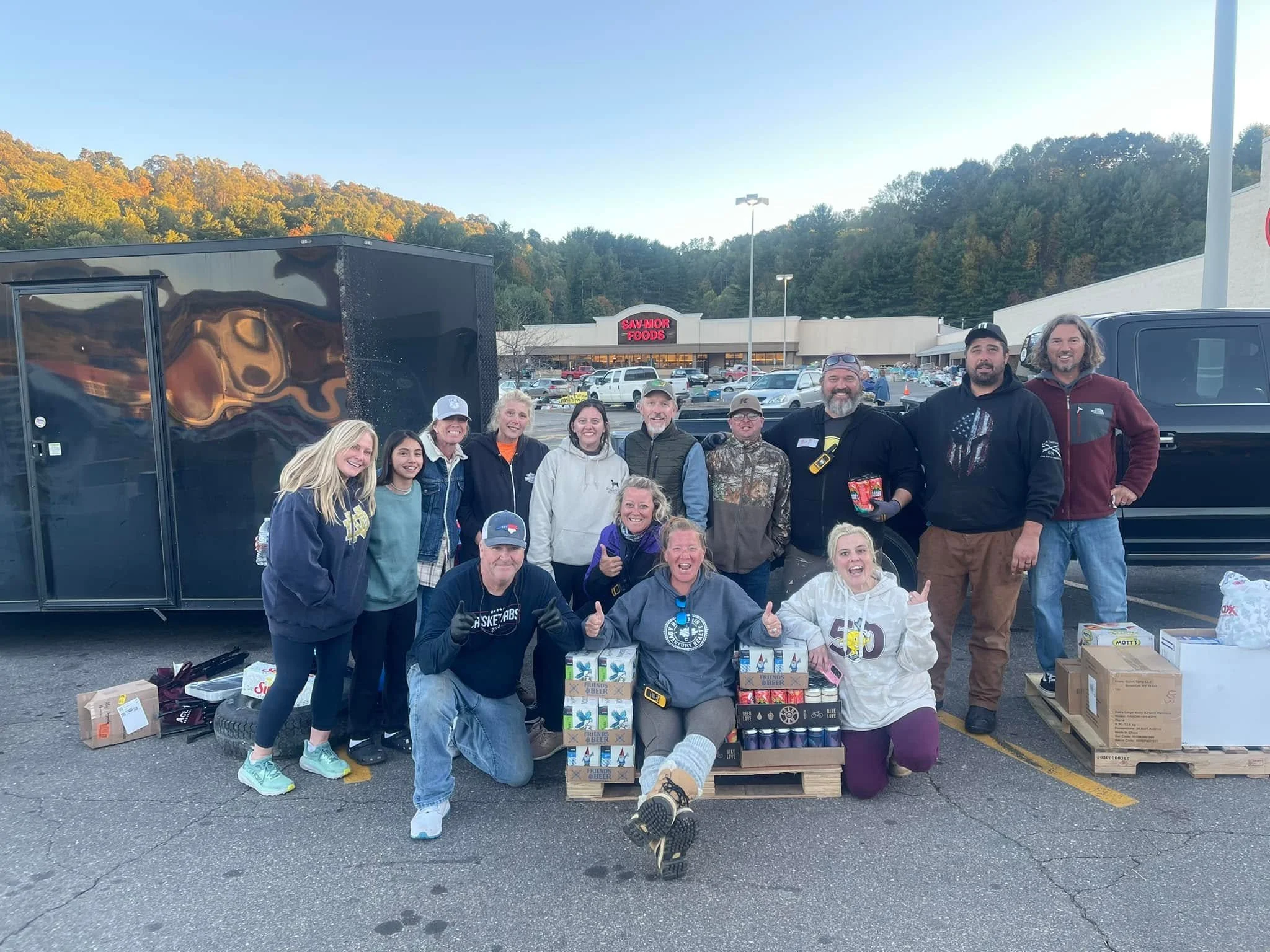 Group of people gathered in a parking lot with boxes of food, smiling and holding up peace signs, in front of a store called Sav-Mor Foods.