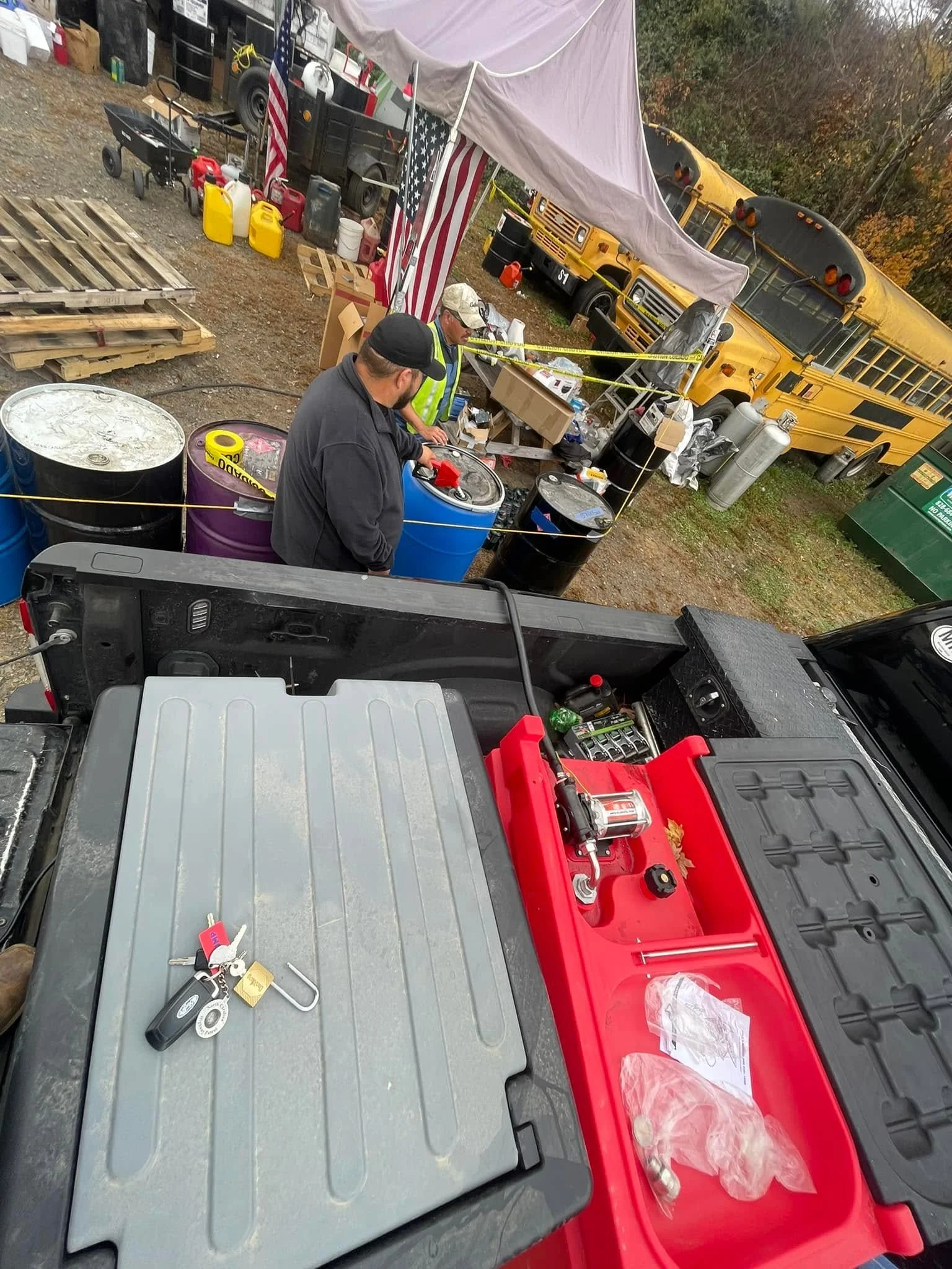 An outdoor scene with a tent, two men, and emergency vehicles. The men are near containers and equipment, with yellow caution tape cordoning off the area. There's a school bus and a utility vehicle in the background.