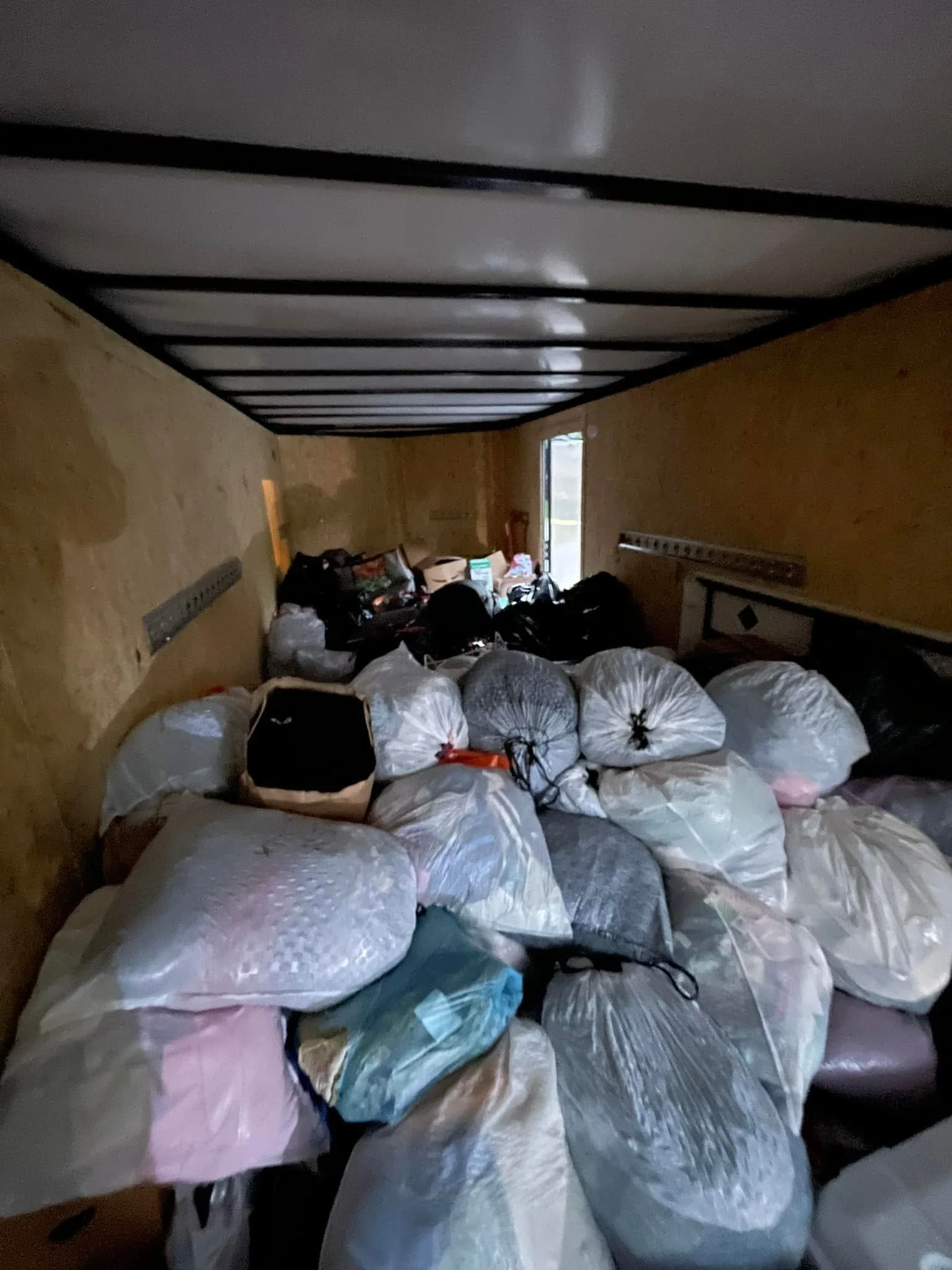 A dimly lit room filled with numerous plastic bags, mostly white and some in colors, piled on the floor. There are some boxes and clutter in the background near a small door or window.