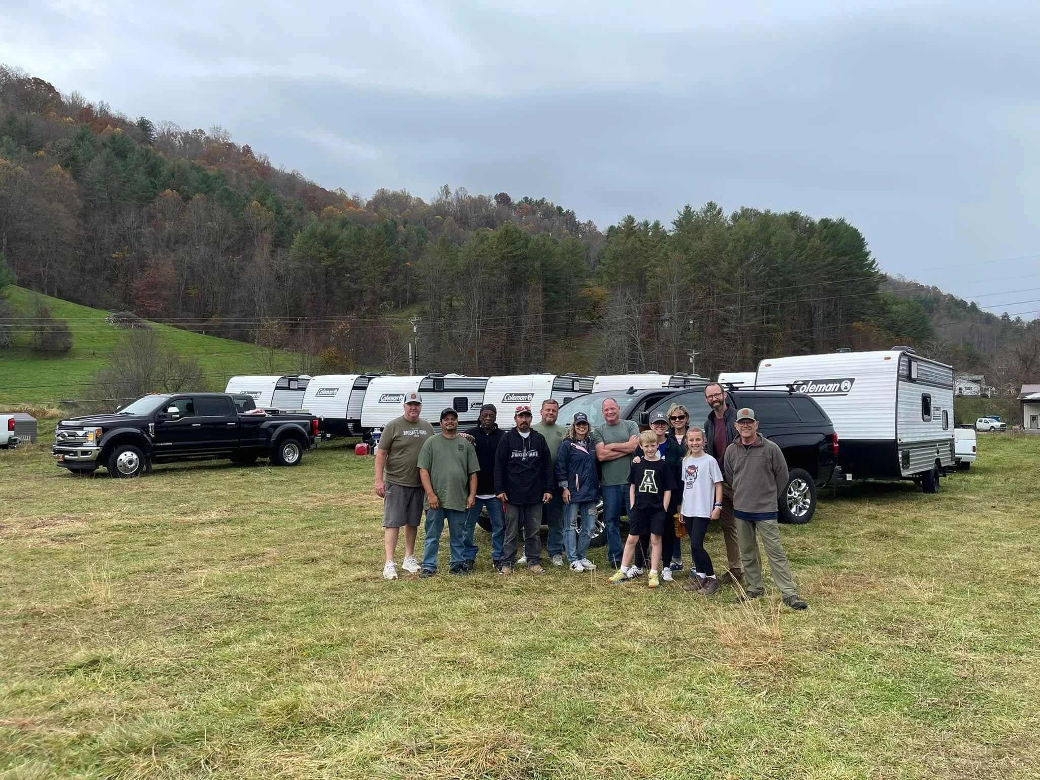 A group of people standing outdoors in front of several travel trailers, with a hillside covered in trees in the background.
