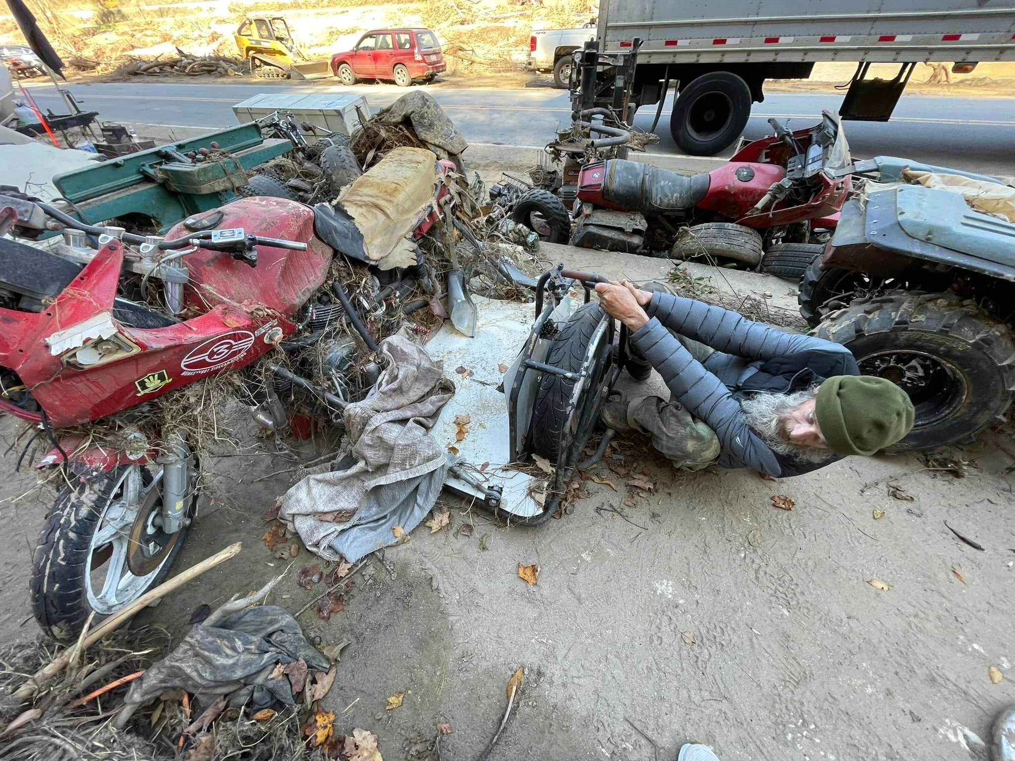 A crash scene showing multiple damaged motorcycles and debris on the side of the road, with a person lying on the ground nearby and a large truck passing by.