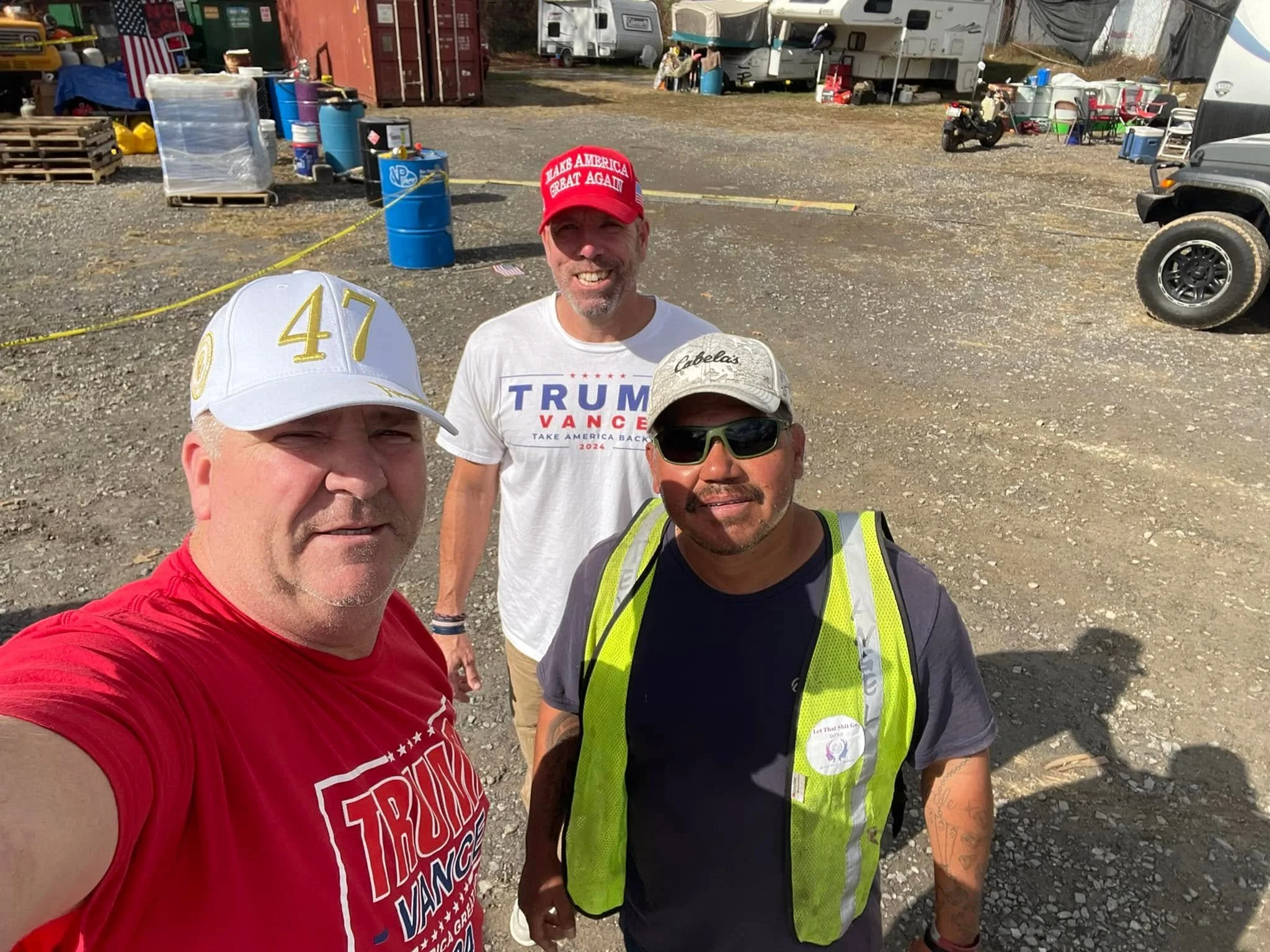 A group of four men taking a selfie outdoors at a gathering or protest. The man in the front is wearing a red T-shirt and a white hat with yellow numbers, while the man to his left is wearing a bright yellow safety vest and a black T-shirt. The man b