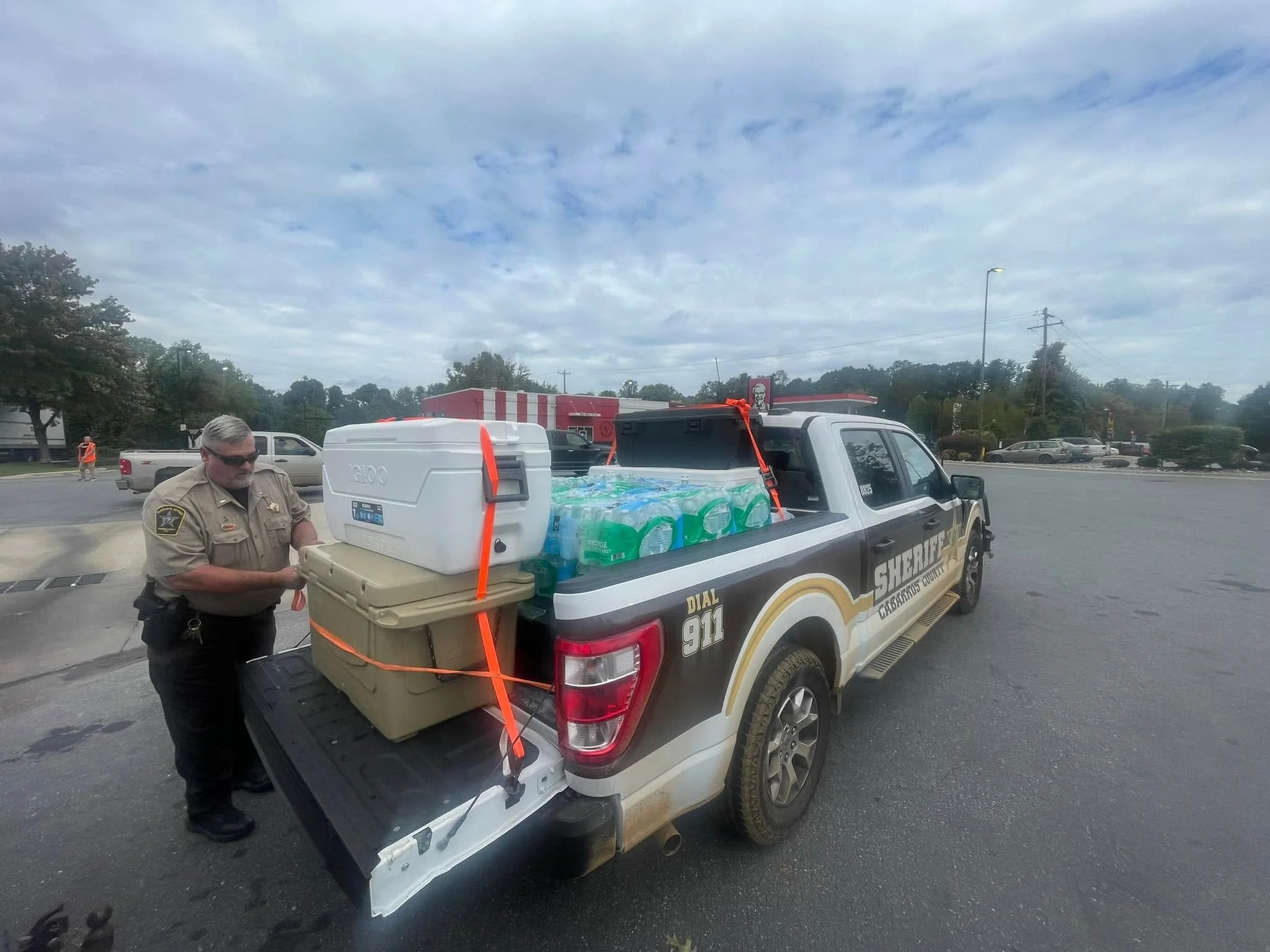 A sheriff's pickup truck with supplies in the bed, including bottled water, a large cooler, and containers, is parked in a lot near a KFC restaurant. A sheriff's officer is standing beside the truck.