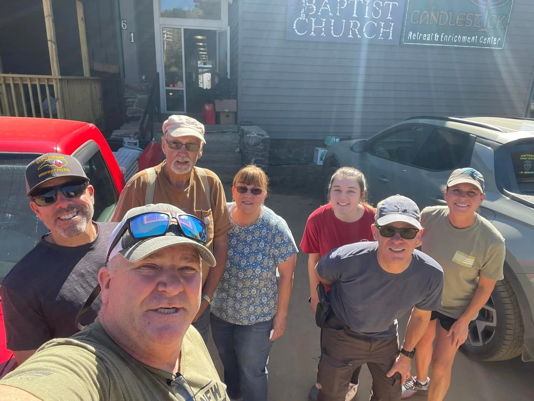 A group of nine people taking a selfie outdoors in front of a building with signs for a church and retreat center. The group includes men and women, some wearing sunglasses and hats.