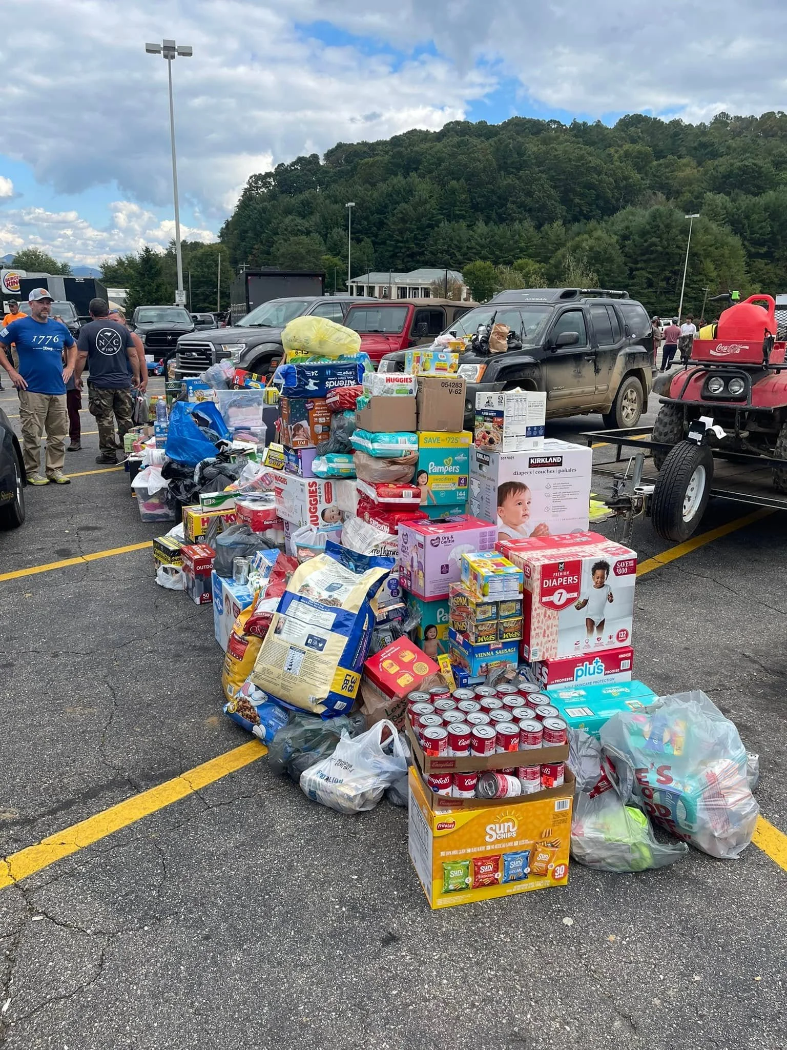 A parking lot filled with various food and supplies, including boxes of diapers, canned goods, snacks, and bottled water, with people gathered around and mountains in the background.