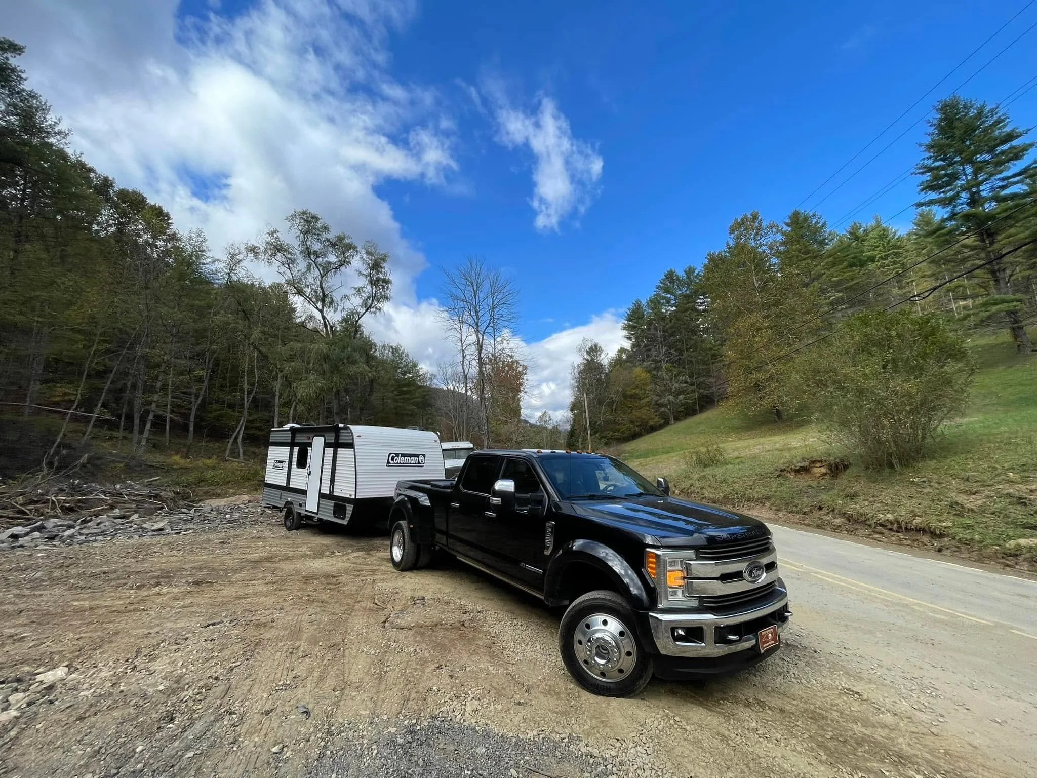 A black pickup truck towing a small white camper trailer on a gravel area beside a two-lane road, surrounded by trees under a blue sky with clouds.