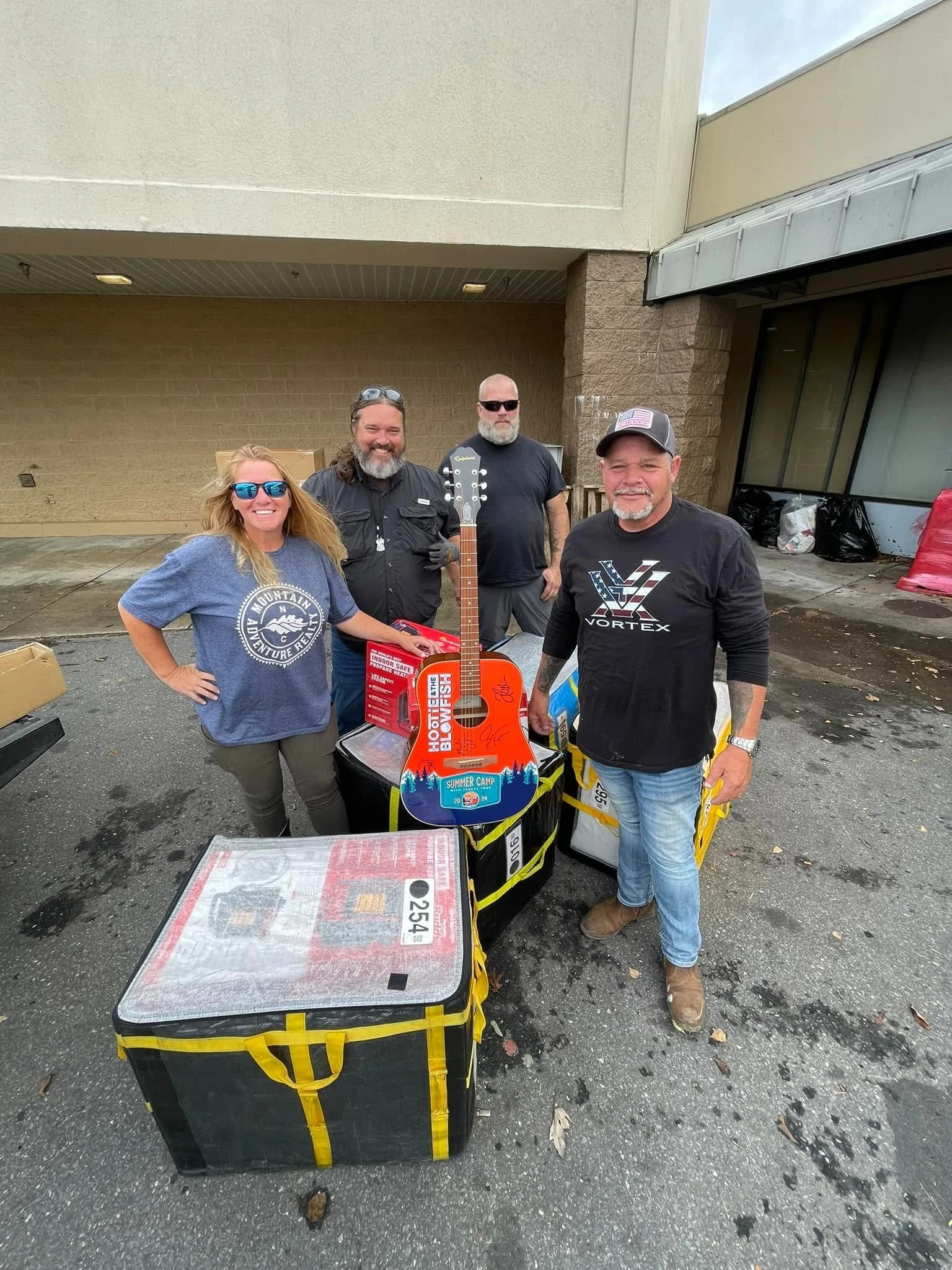 Four people standing outdoors in front of a building, with supplies such as a large emergency kit, a guitar, and various bags around them, smiling and posing for the photo.