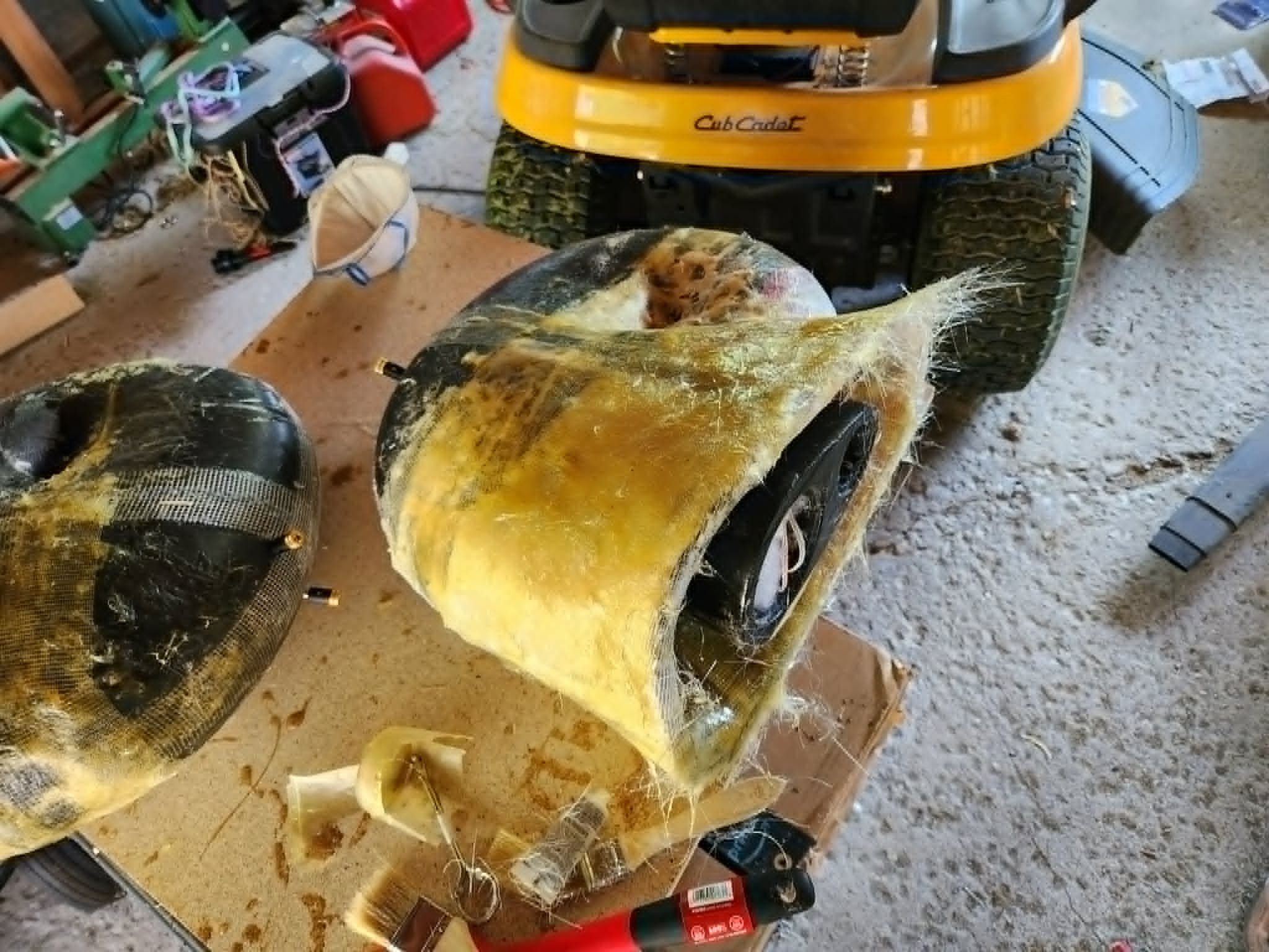 A workbench in a garage with a large yellow lawn tractor with a broken or damaged body, and parts of the body removed exposing internal components, with tools and miscellaneous items scattered around.