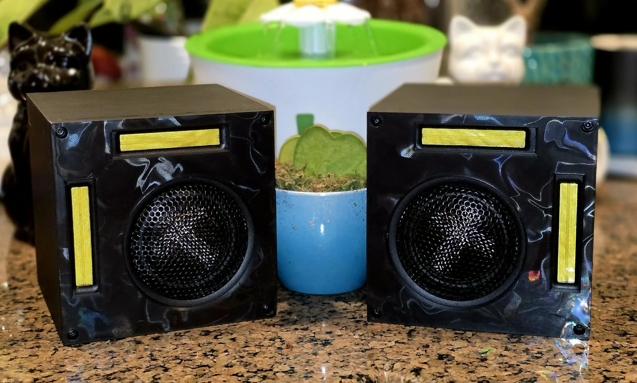 A pair of black speakers with yellow accents placed on a speckled countertop. Behind the speakers, there is a small green and white humidifier, with various decorative items including a black bear figure, a ceramic cat, and jars in the background.