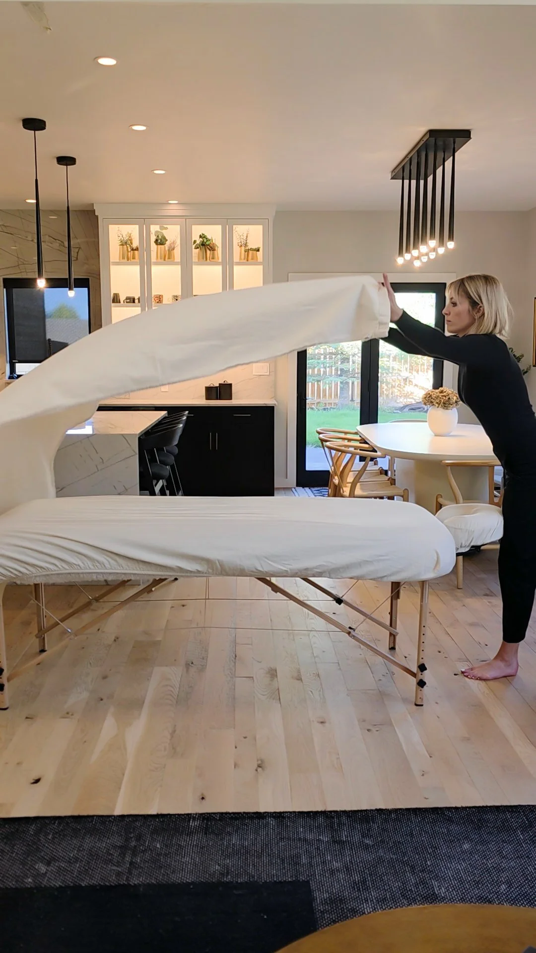 A woman adjusts a white massage table in a kitchen and dining area of a modern home, with a person lying on the table and a large window showing an outdoor yard.