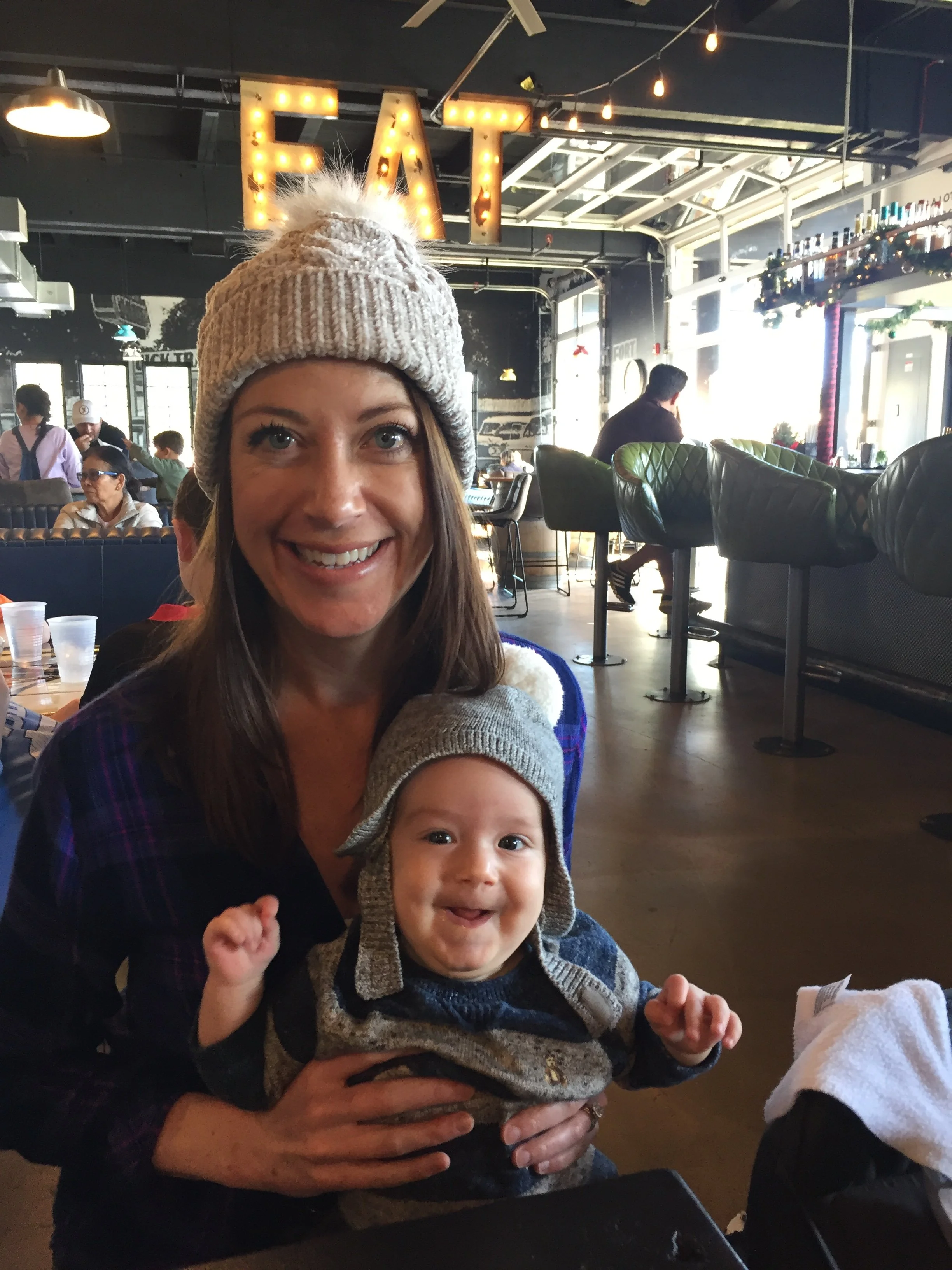 Sheena Nageli. A woman with long brown hair wearing a beige knitted hat, smiling, holding a baby in a dark sweater and beige hat, sitting at a restaurant with other patrons in the background.