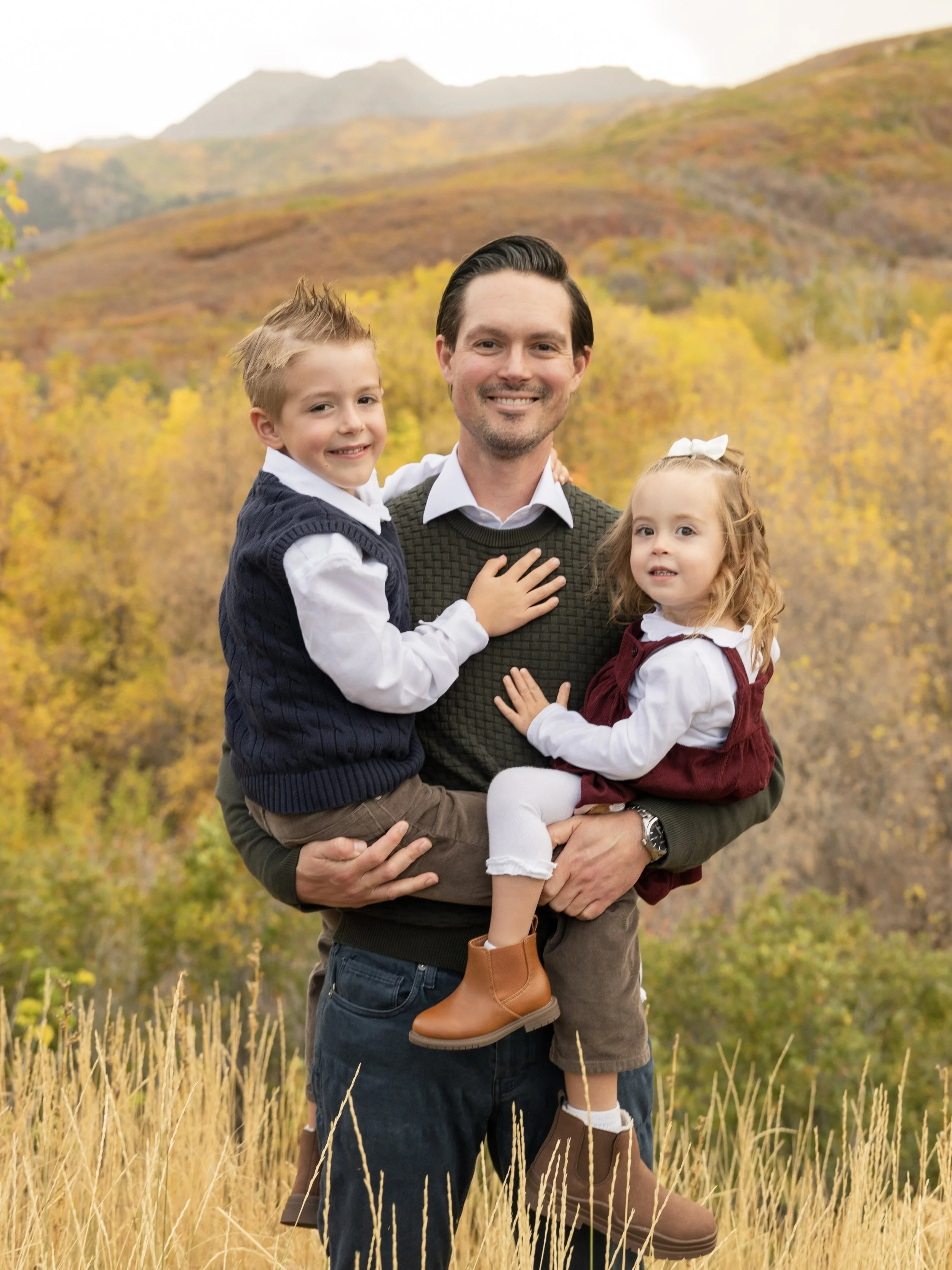 Nick Nageli.  Nicholas Nageli. A man holding two children in a field of tall yellow grass with autumn trees and mountains in the background.