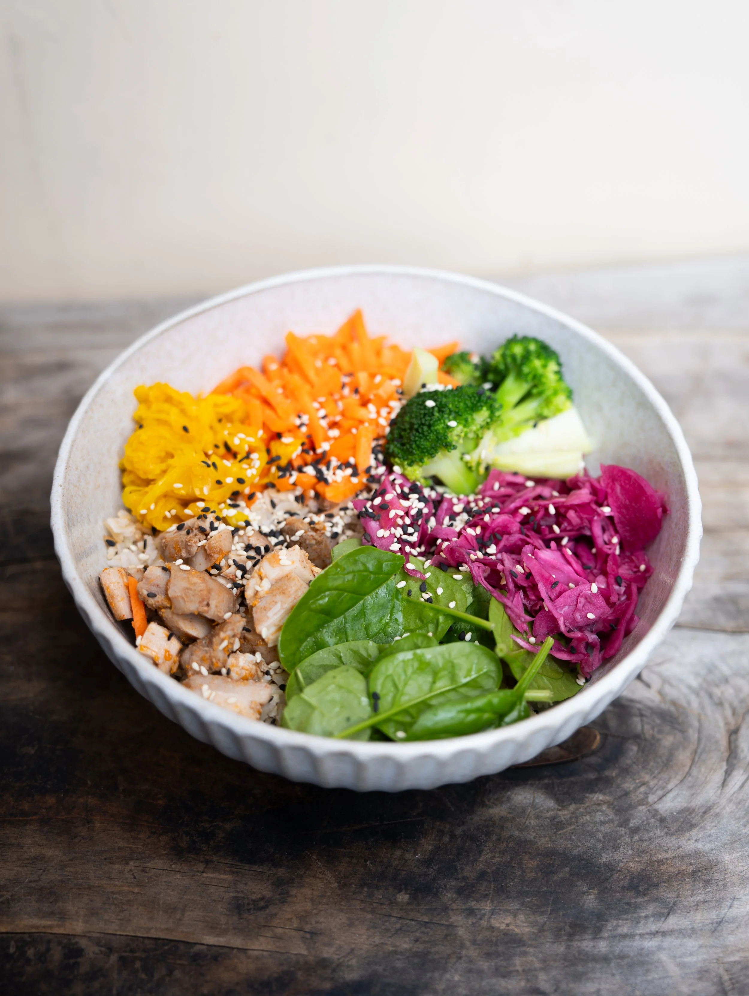 A bowl of colorful Asian-style salad with vegetables such as broccoli, red cabbage, carrots, yellow pickled vegetables, and green leafy vegetables, topped with sesame seeds.