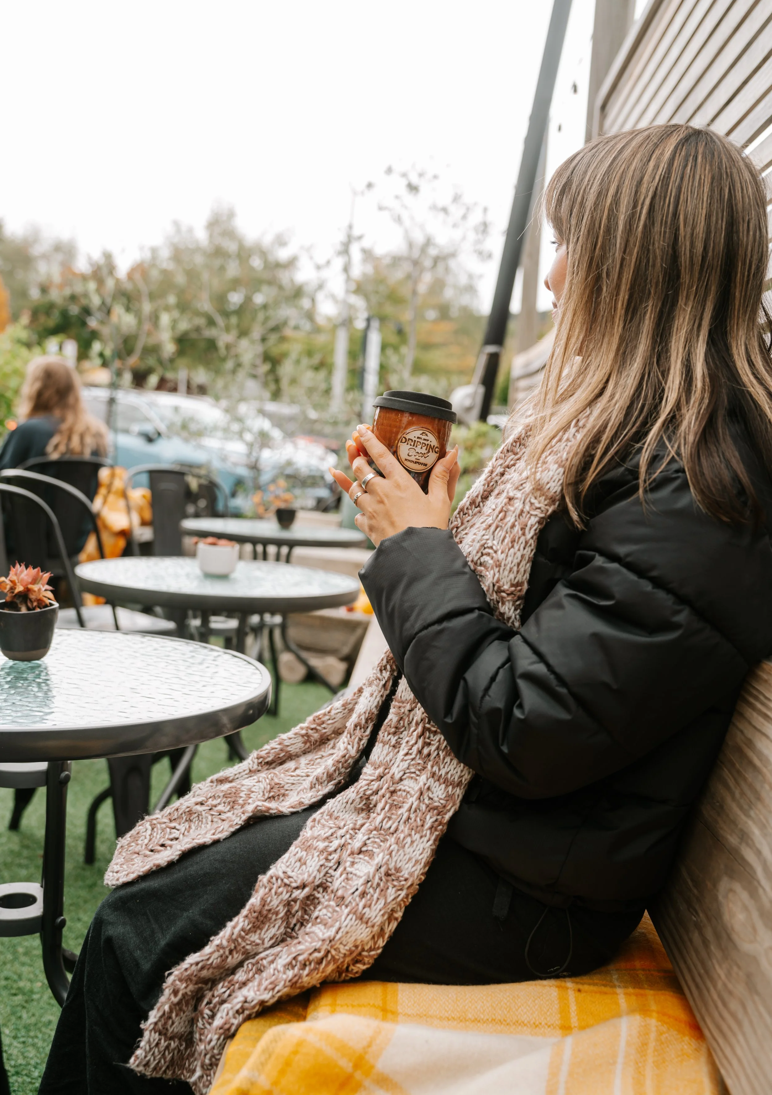 A woman with light brown hair sitting outdoors, holding a coffee cup, wearing a black jacket and a pink scarf. There are tables with potted plants and other people in the background.