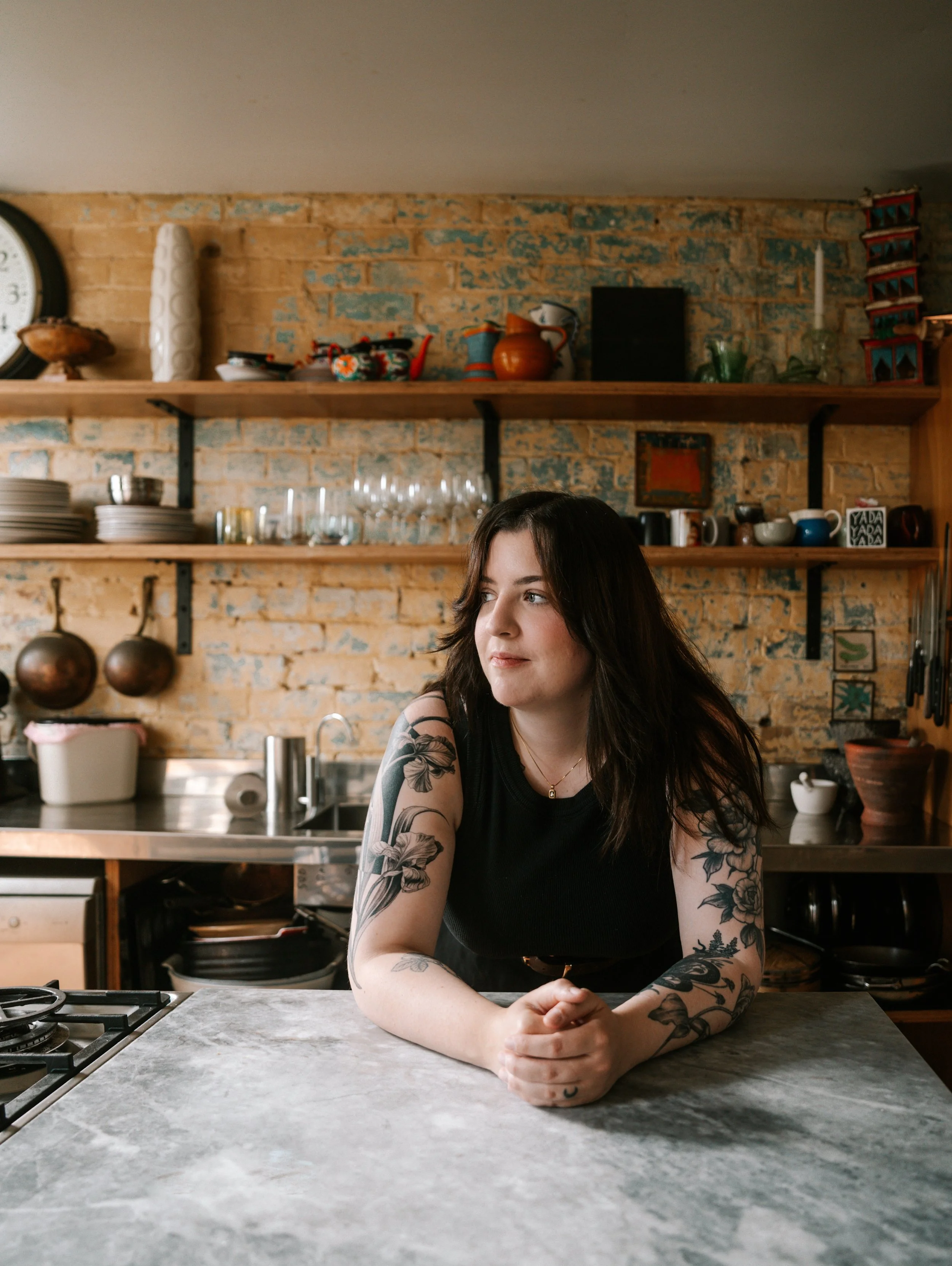 A woman with tattoos on her arms sitting at a kitchen counter, looking to the side, with a rustic brick wall with shelves and decorative items behind her.