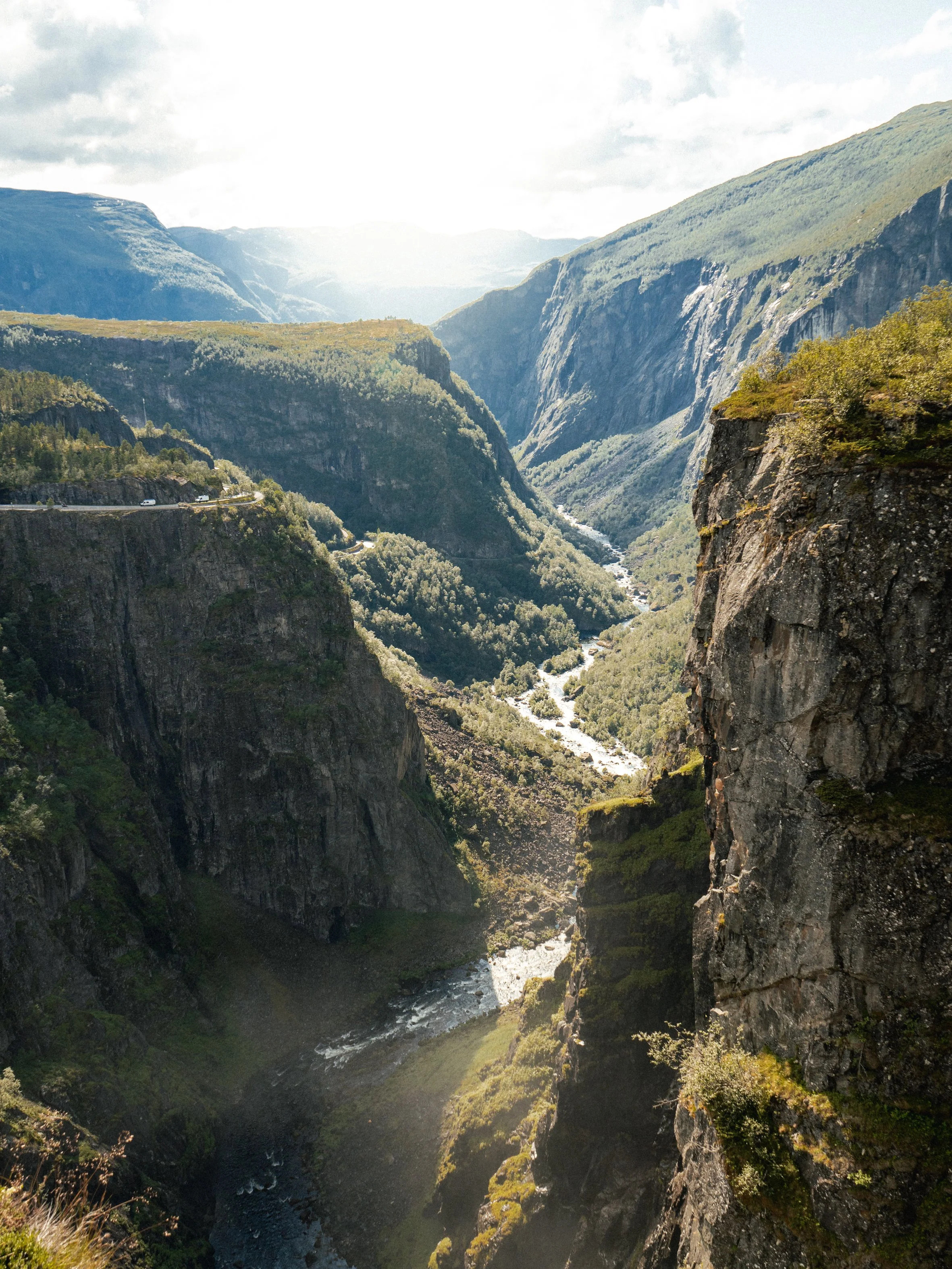 A deep canyon with steep rocky cliffs on both sides, a river flowing through the bottom, and mountainous terrain in the background. Bright sunlight illuminates the scene, highlighting the lush greenery on the cliffs and the river below.