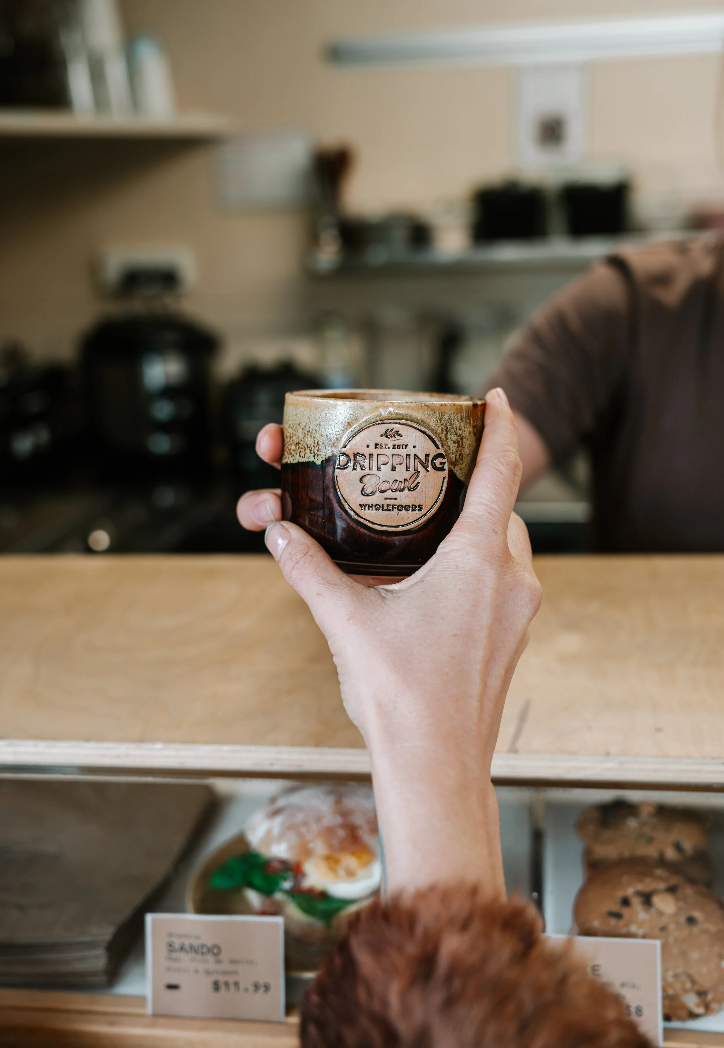 A hand holding a ceramic mug with the words 'Dripping Bowl' printed on it, in a bakery or cafe setting. In the background, there are baked goods on display and a blurred person with brown hair.