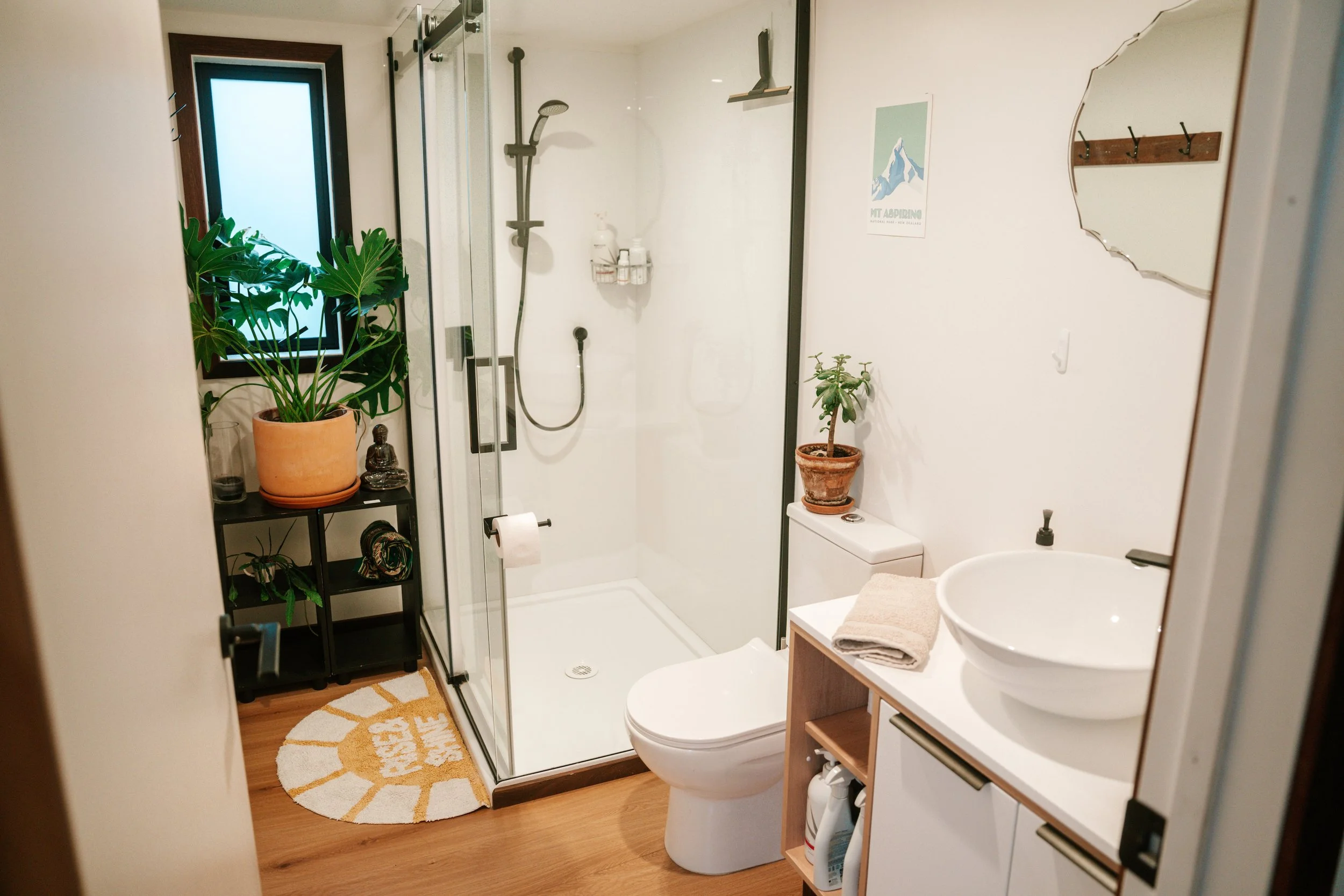 A small bathroom with a glass shower enclosure, a toilet, a white sink, and various potted plants, a mirror, and artwork on the walls.