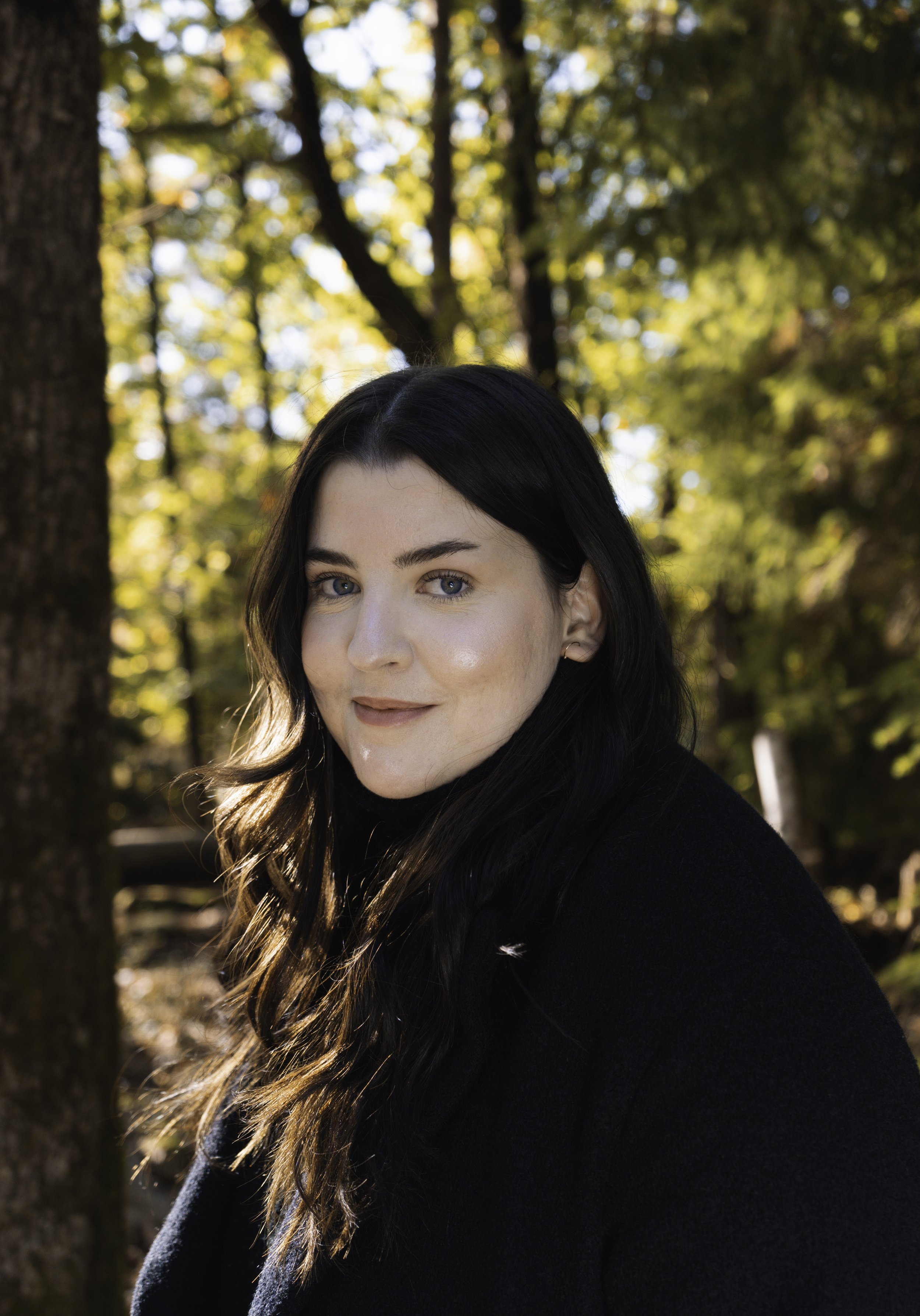 A woman with long dark hair and blue eyes outdoors in a wooded area with sunlight filtering through the trees.