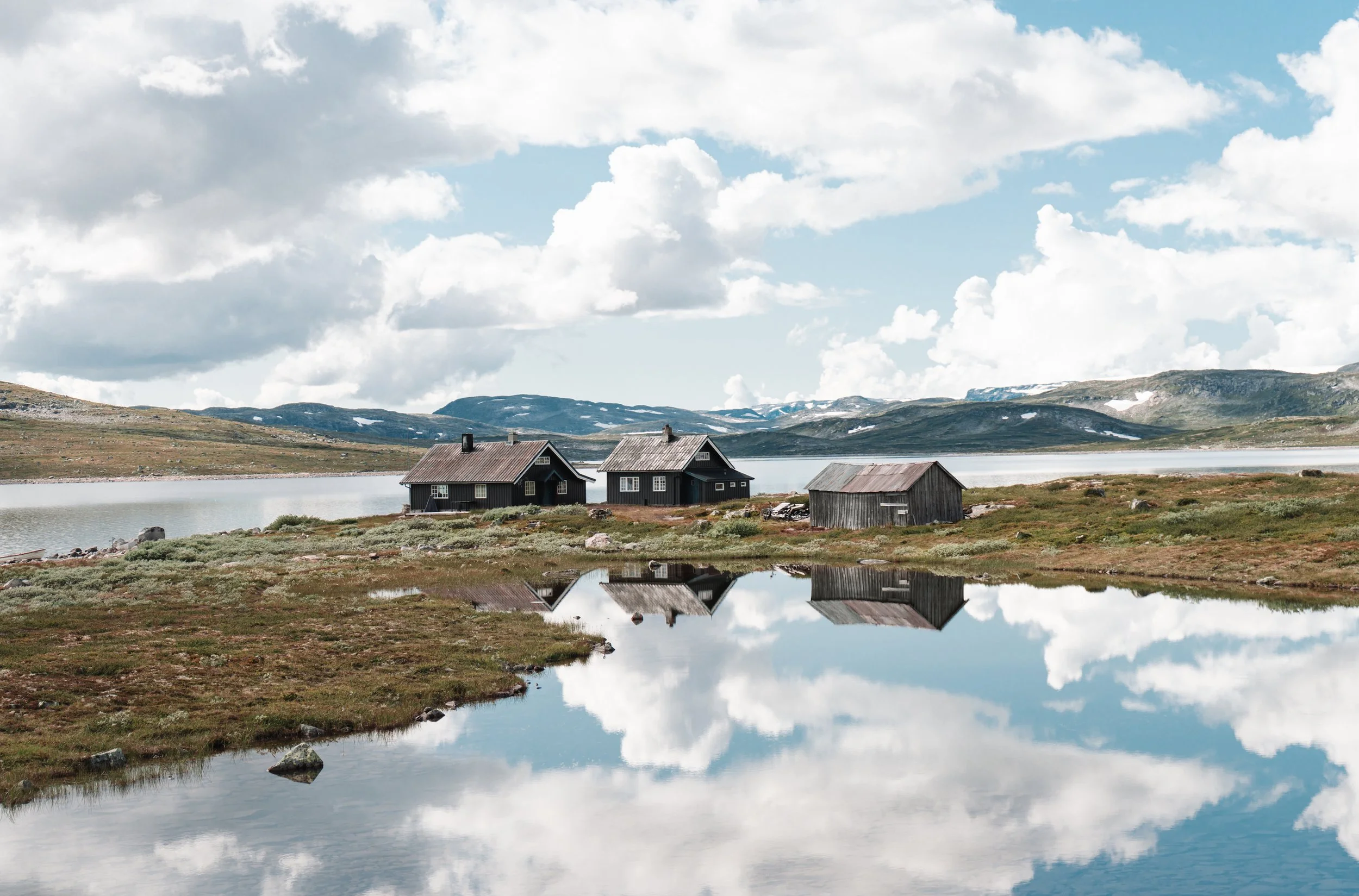 Scenic view of a lake with clear reflections of the sky, clouds, and small wooden houses on the shoreline, set against a backdrop of distant mountains and a partly cloudy sky.