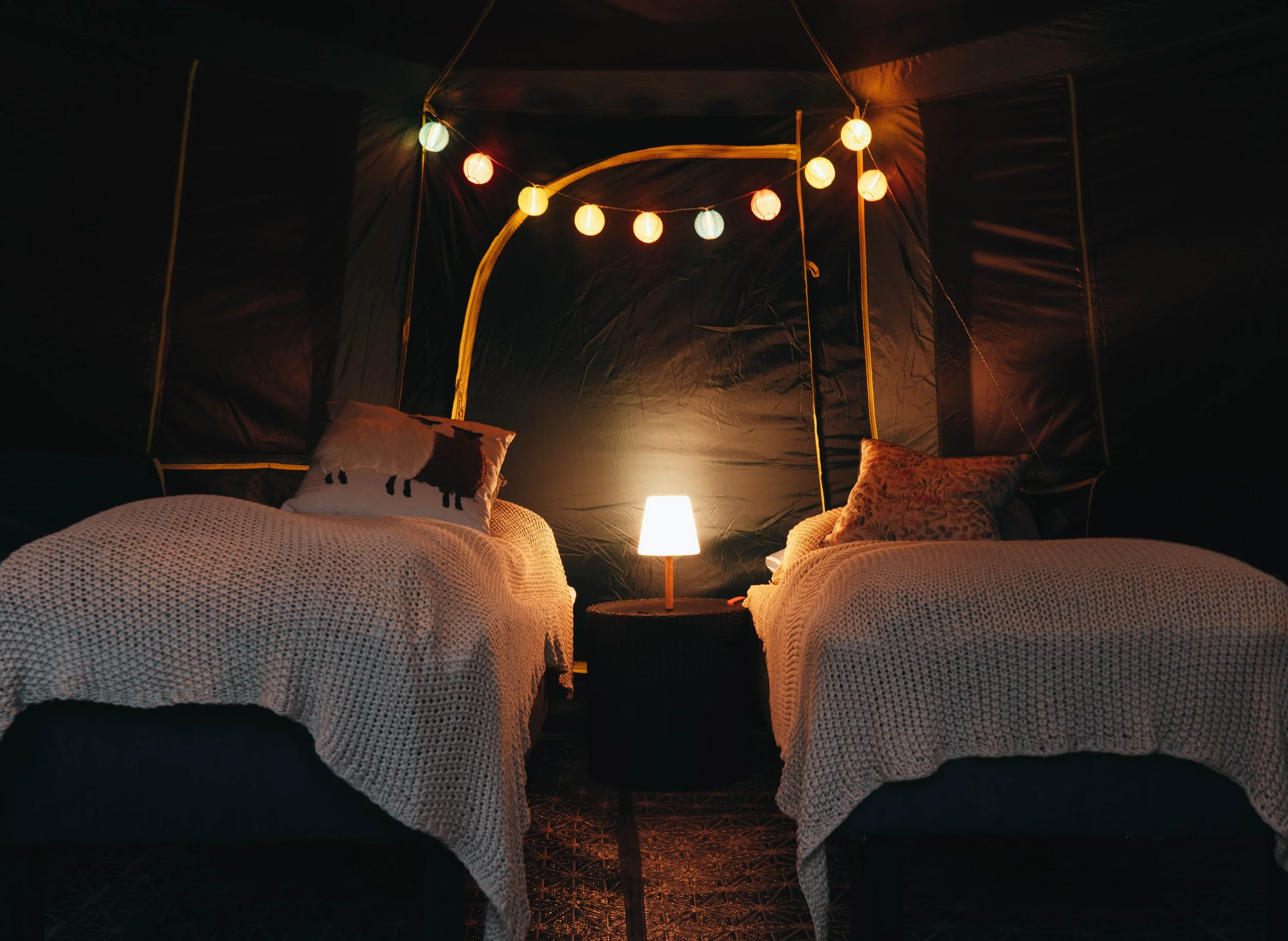 Dimly lit bedroom with two beds, a small table in between, a lamp, string of colorful lantern lights on the wall, a pillow with a llama design on one bed, and a patterned rug on the floor.