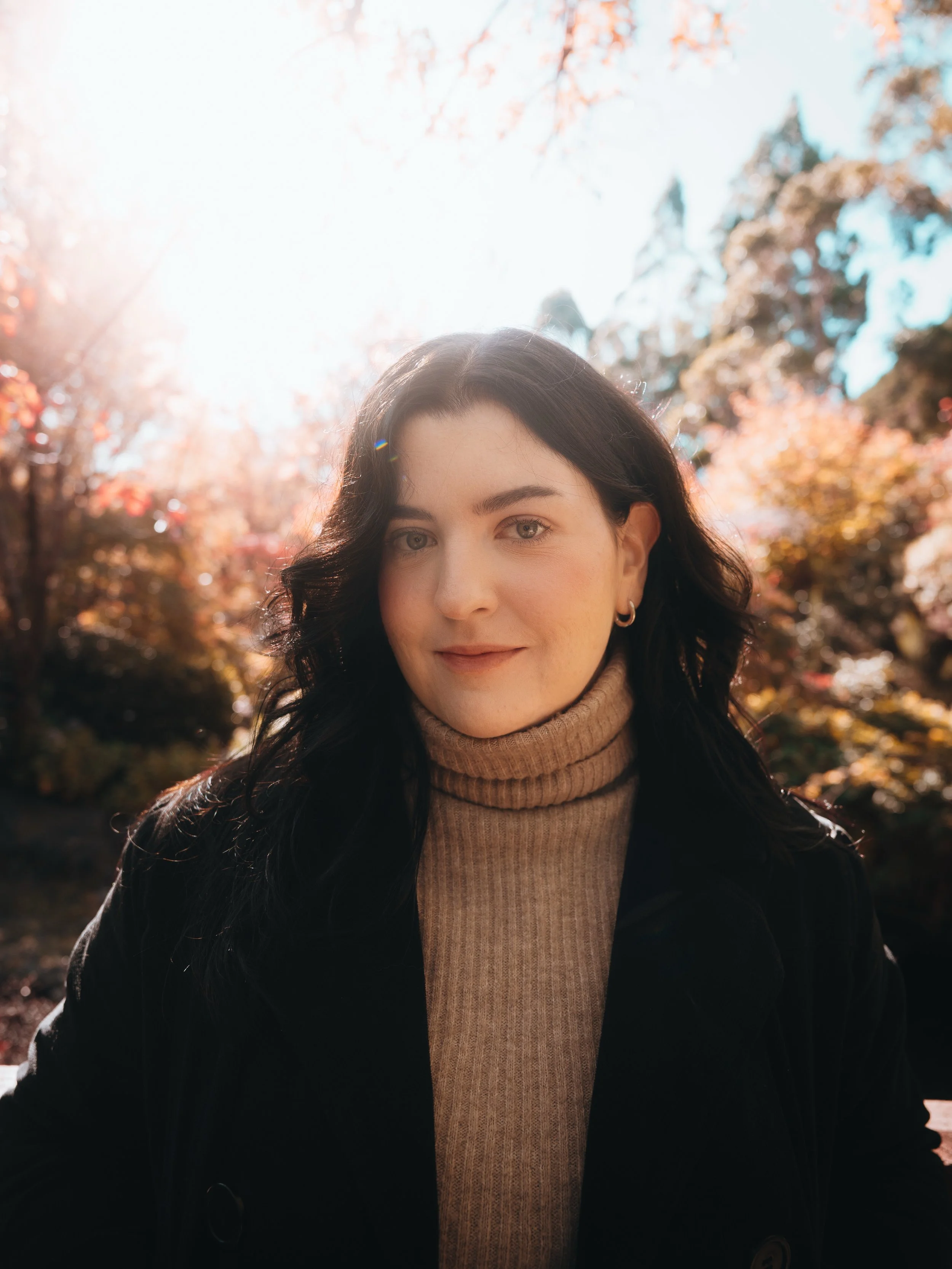A woman with dark hair, light-colored eyes, wearing a beige turtleneck sweater and black coat, standing outdoors with autumn-colored trees in the background, sun shining brightly.