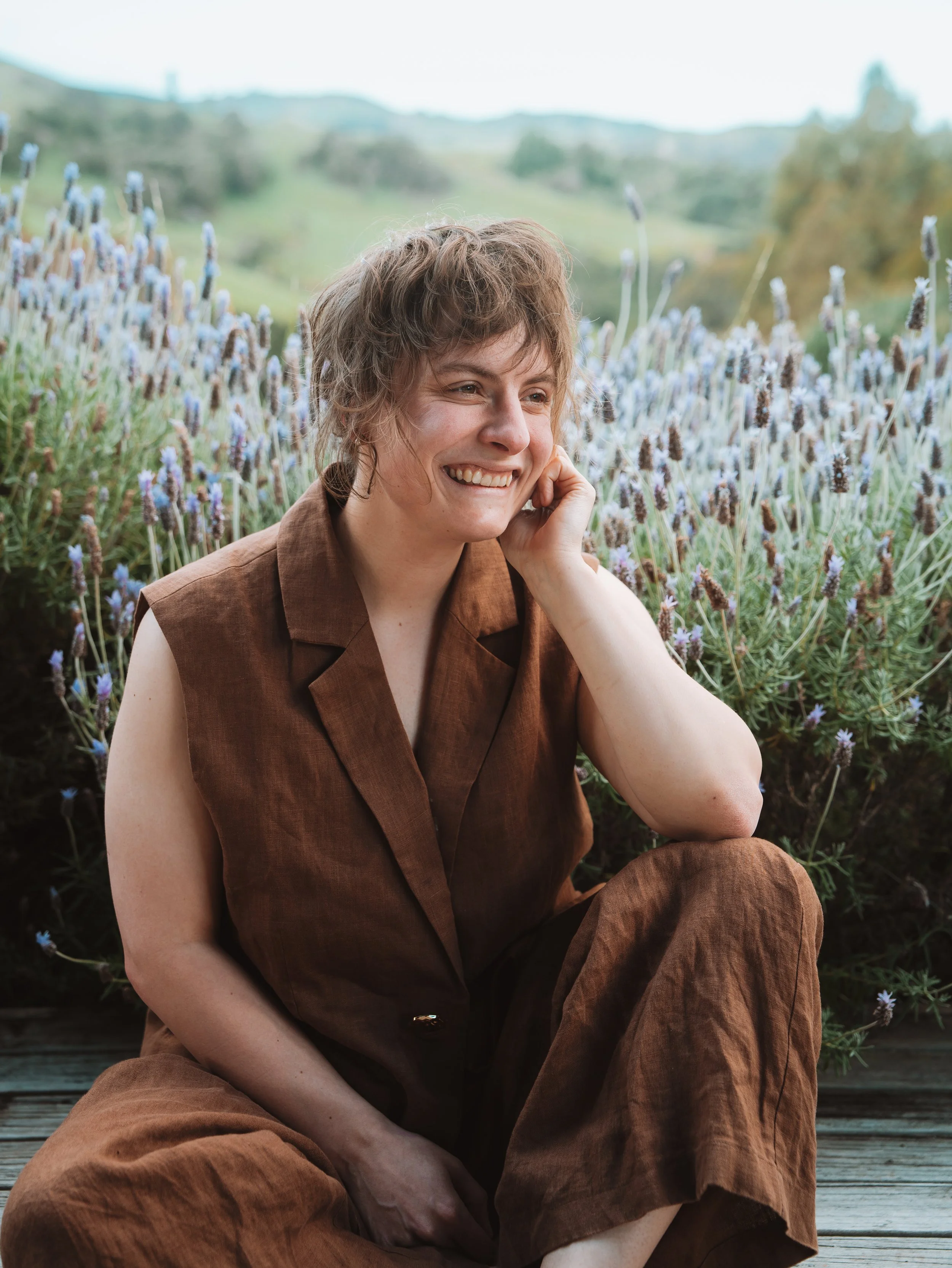 Smiling woman sitting outdoors among lavender flowers, with a green hilly landscape in the background.