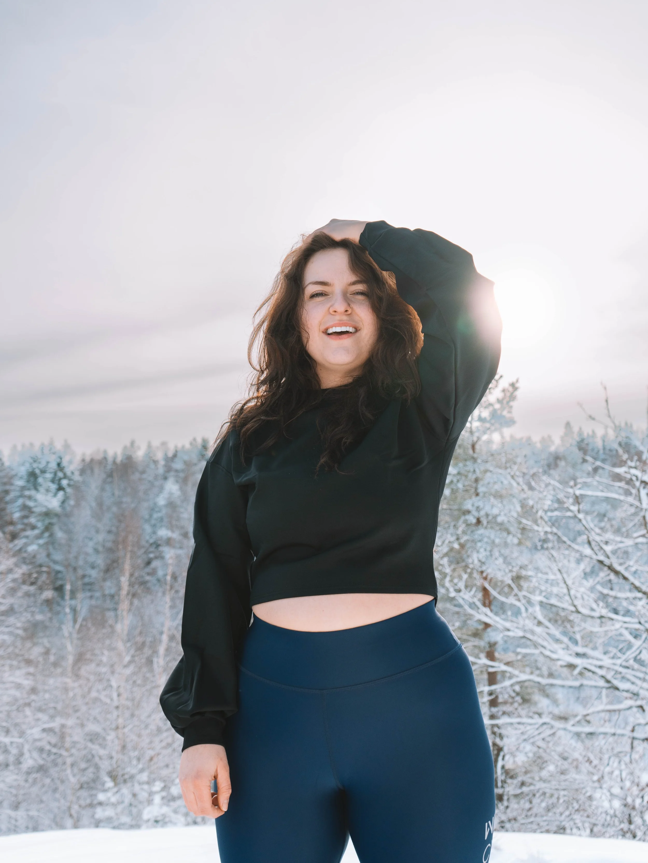 Young woman with long curly hair smiling outdoors in winter, wearing a black long-sleeve top and navy leggings, with snow-covered trees in the background.