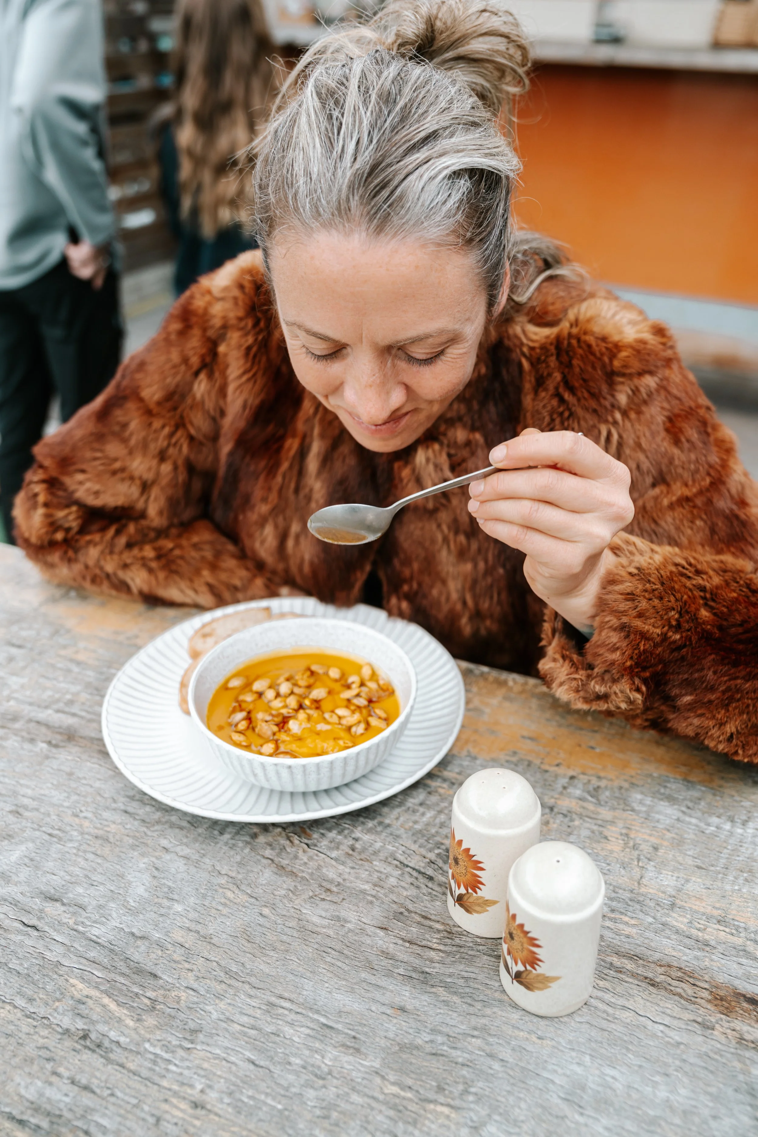 A woman with gray hair wearing a fur coat is sitting at a rustic wooden table, smiling, while holding a spoon with some kind of soup or pudding. The bowl in front of her contains a yellowish liquid with beans or chickpeas. Two white salt and pepper s