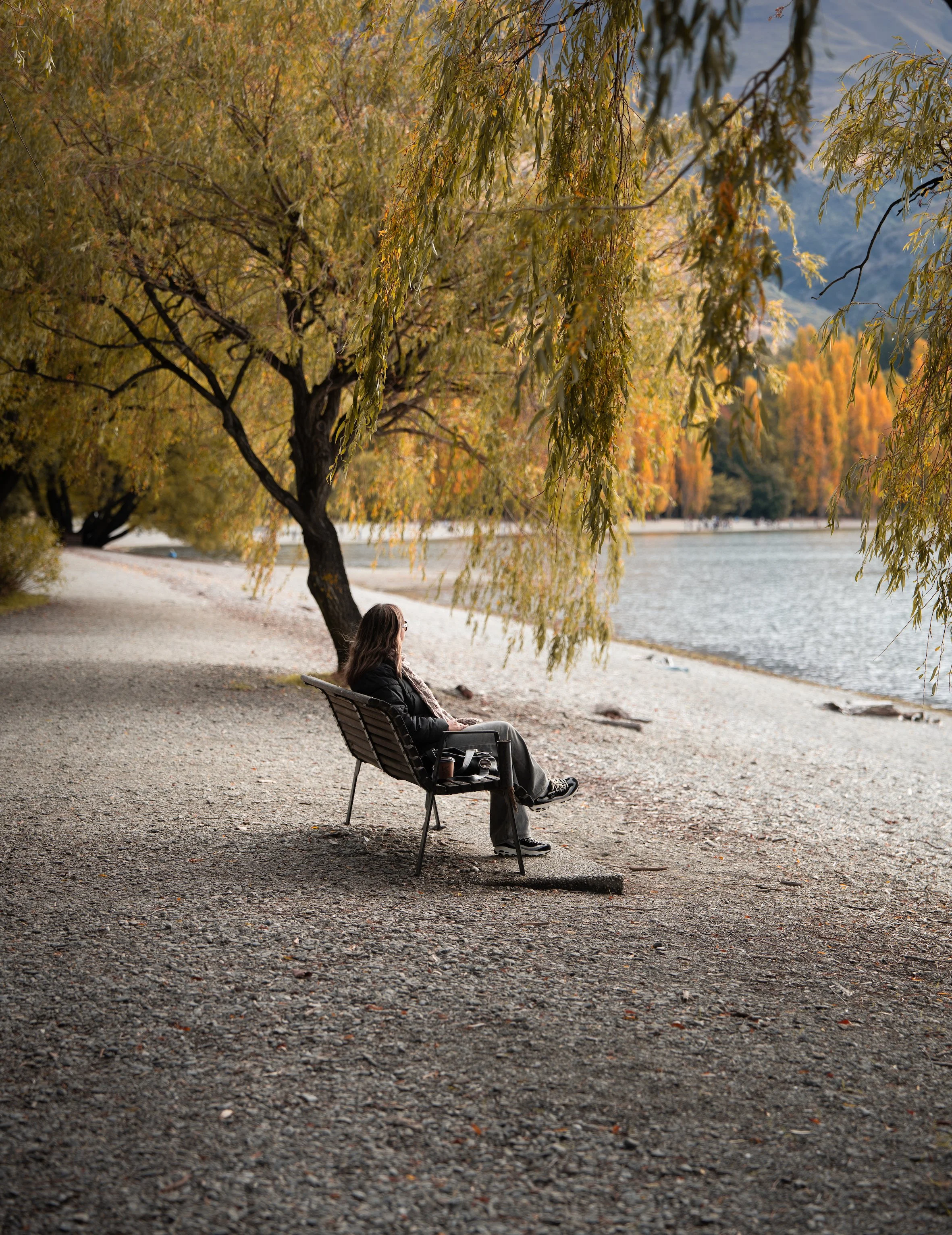 A woman sitting on a bench under a tree by a lake during fall, with colorful foliage.